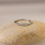 The Baguette Diamond Ring features a simple silver band and a rectangular baguette diamond in an east-west bezel setting, showcased on a smooth, light-colored stone.