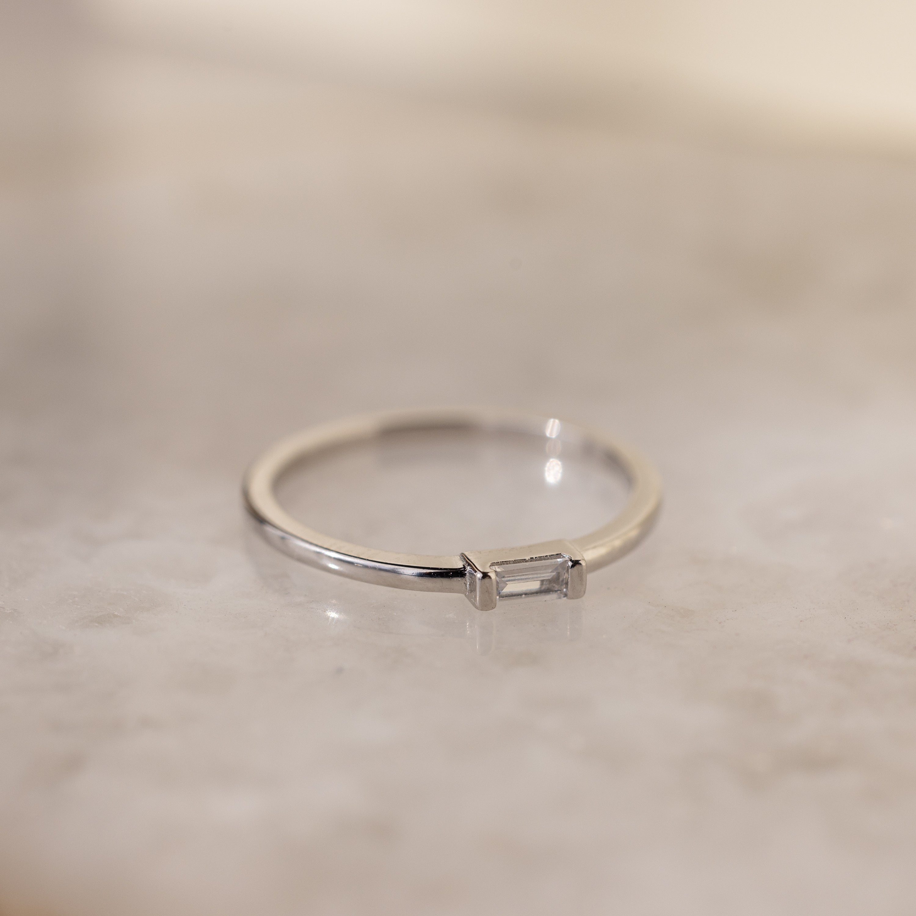 The Baguette Diamond Ring in Silver showcases a dainty baguette diamond set east-west in an elegant bezel setting, displayed on a light marbled surface.