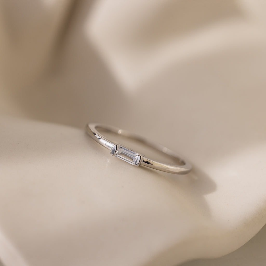 The Baguette Diamond Ring in Silver features a small rectangular stone set east-west in a delicate bezel, showcased on a soft, cream-colored textured surface.