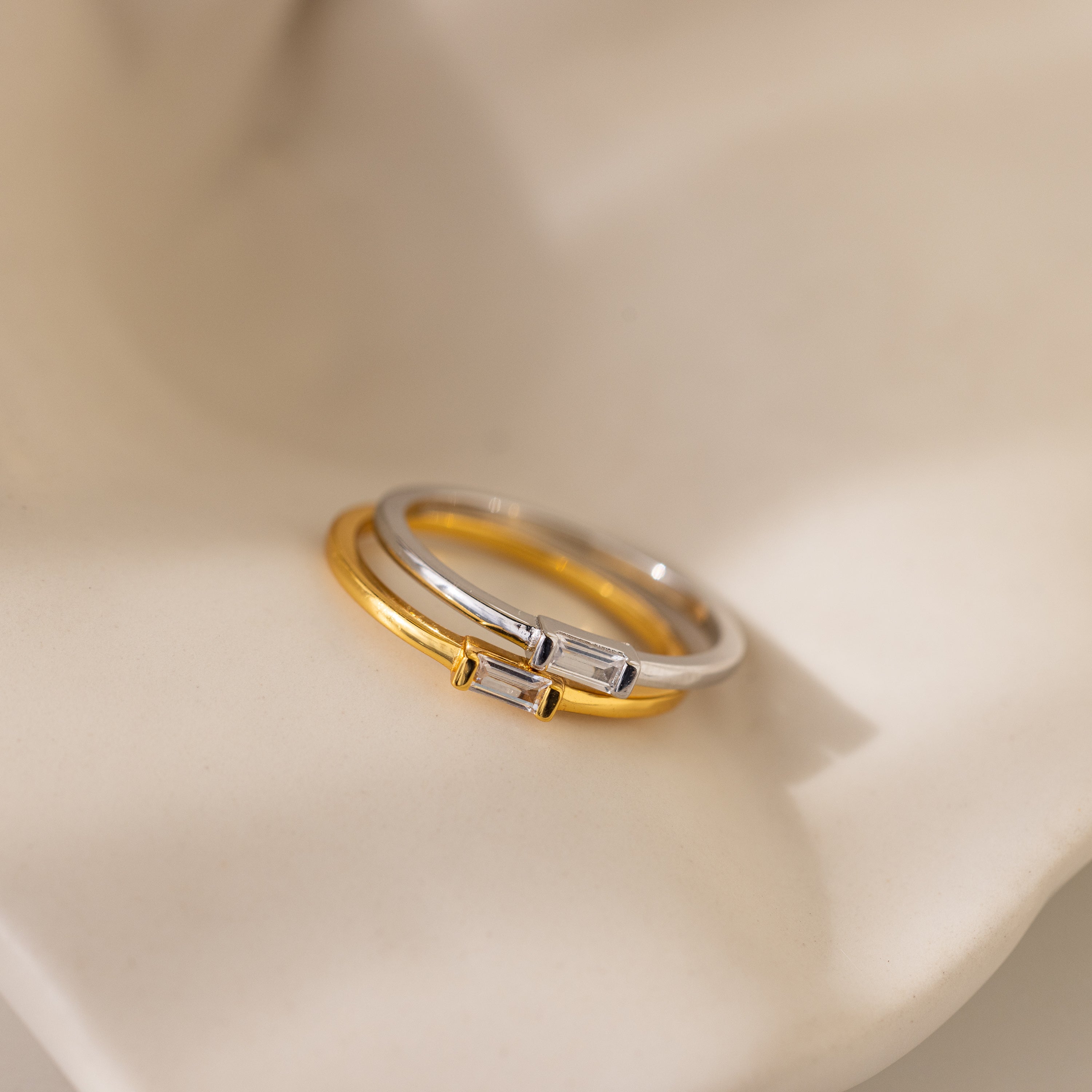 Two Baguette Diamond Rings, one in gold and one in silver, with slim bands and rectangular diamonds set horizontally, are displayed on a soft beige surface.