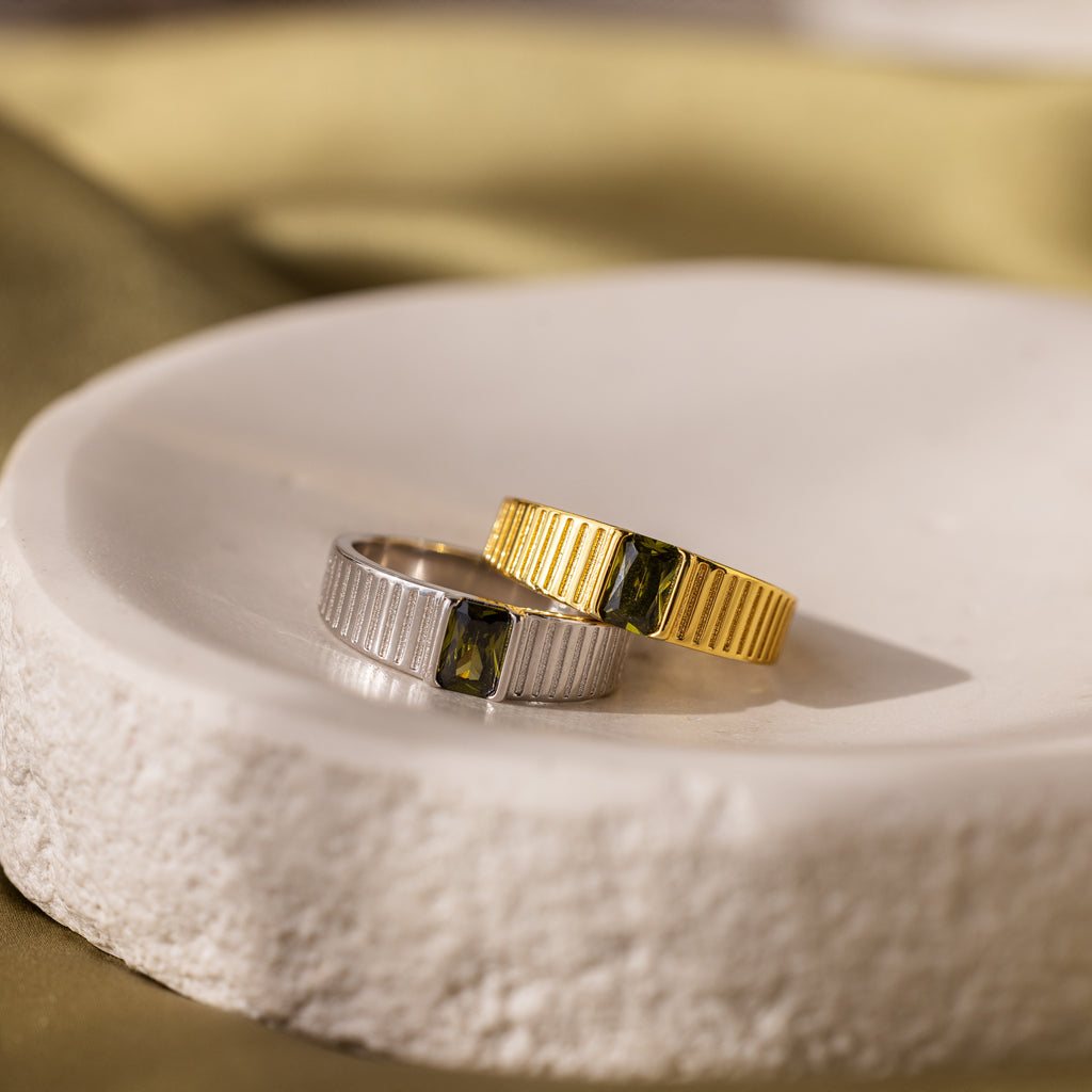 A chunky silver signet ring and the Olive Peridot Signet Ring, both featuring vivid green gemstones, are elegantly displayed on a white ceramic dish.