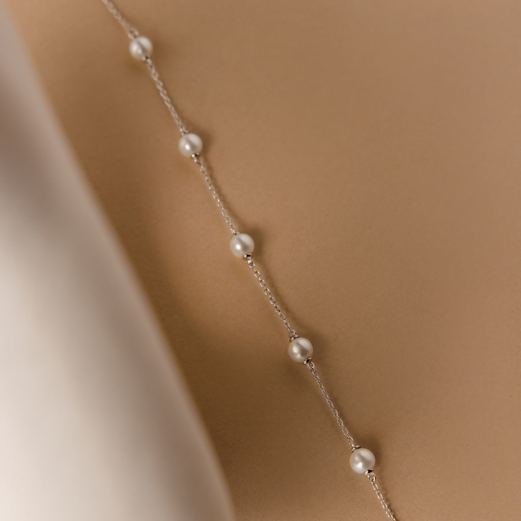 The Pearl Station Bracelet in Sterling Silver features delicate pearls on a fine silver chain, elegantly showcased against a soft beige background.