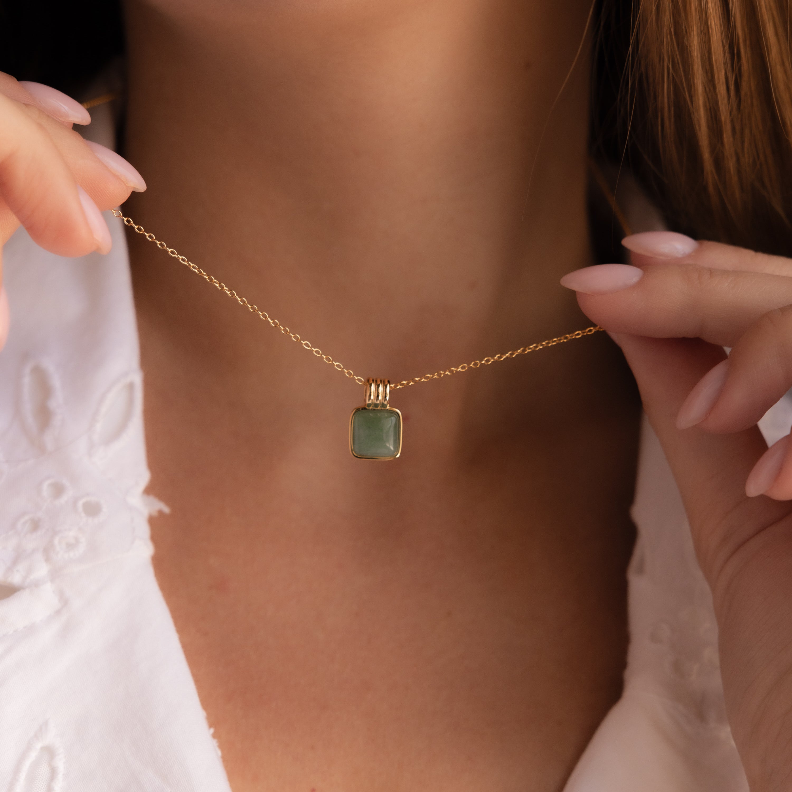 A woman wearing a white embroidered top holds the Jade Pendant Necklace in 18K Gold, featuring a square green jade stone believed to bring luck.