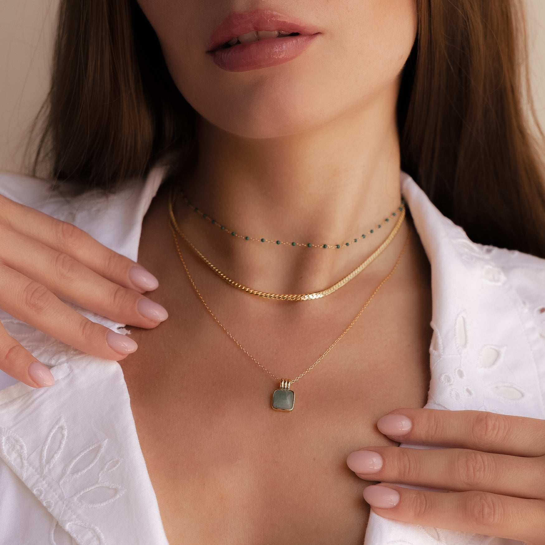 A woman, in a white embroidered shirt, gently touches her collar while wearing the Jade Pendant Necklace with a green jade stone—a symbol of new beginnings.