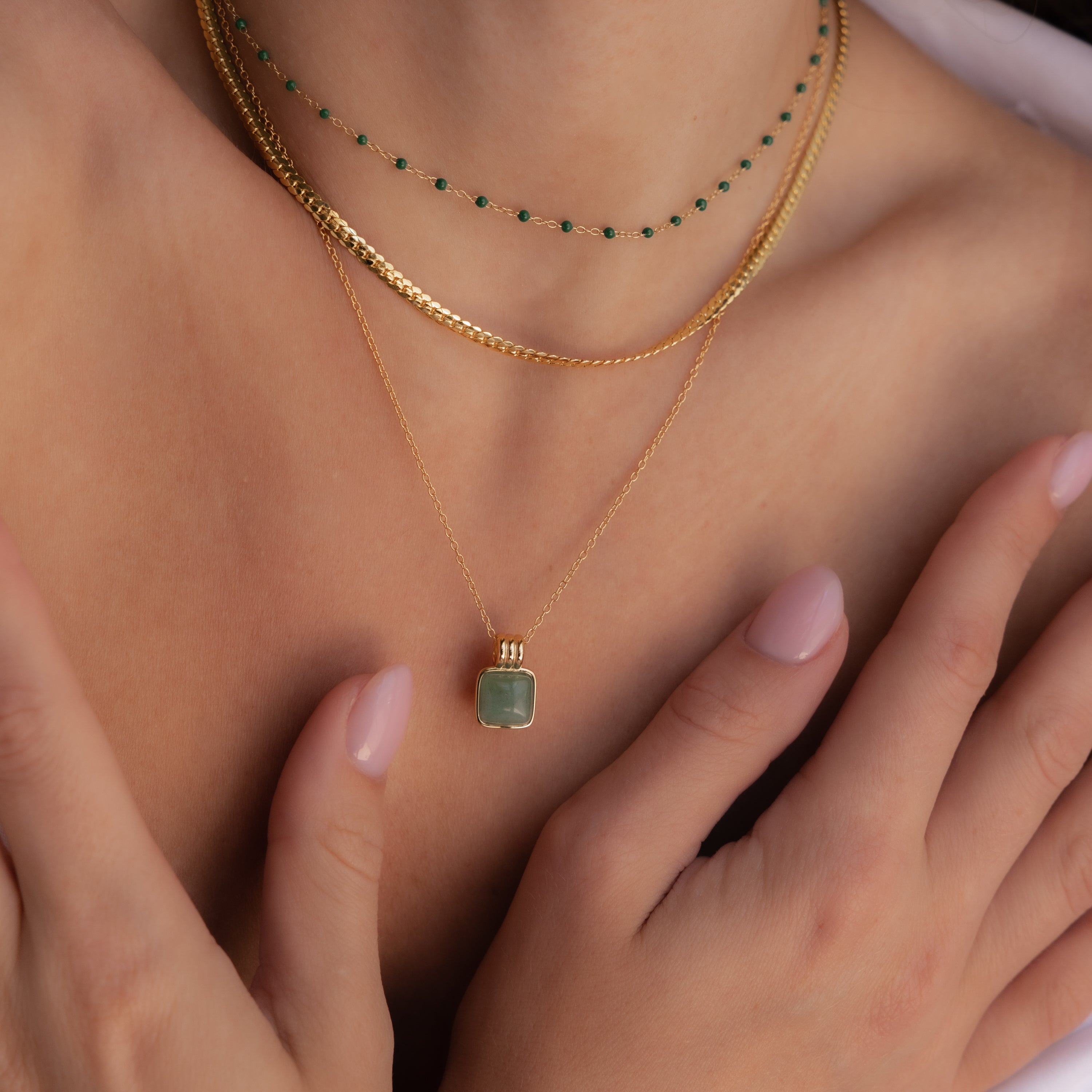A woman with light pink nails rests her hands on her chest, wearing layered gold necklaces featuring the Jade Pendant Necklace, symbolizing new beginnings.