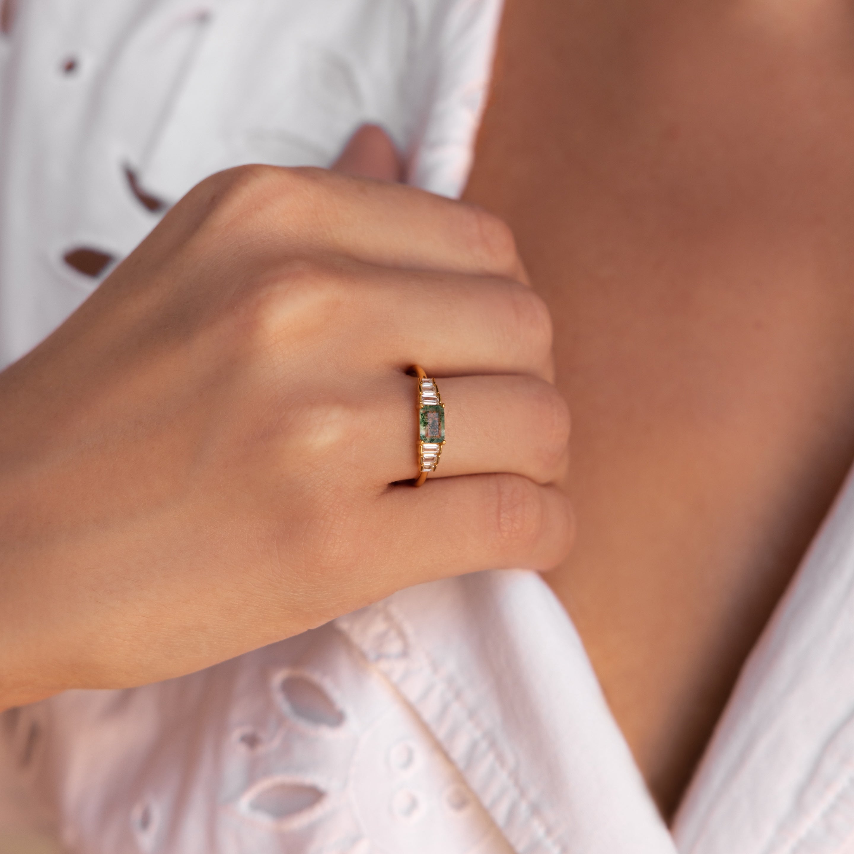 Moss agate gemstone ring with a rectangular green center stone and baguette-cut accents, worn on the ring finger, paired with a white embroidered blouse.