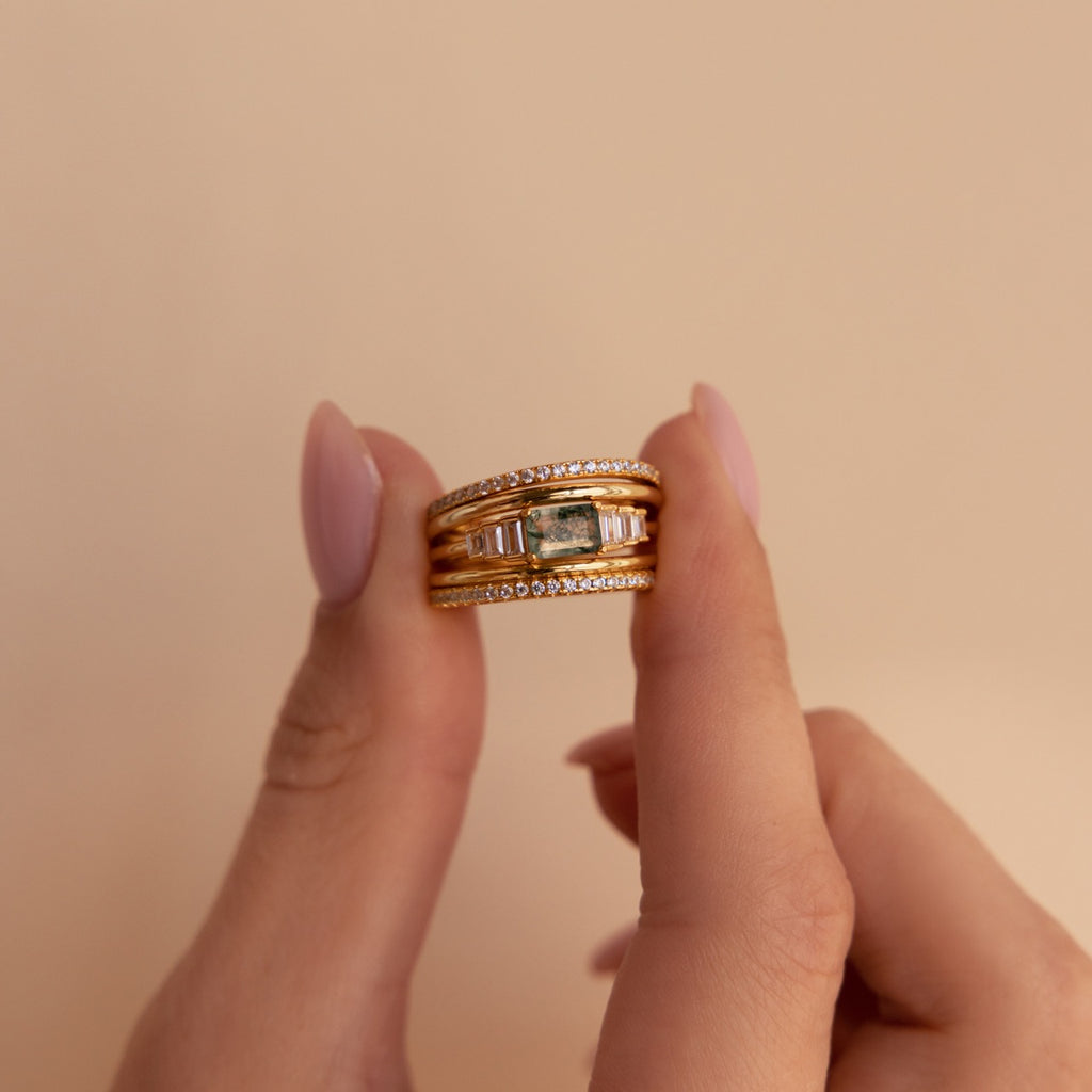 Moss agate ring stacked with three other gold bands, including pavé and domed styles, held between two fingers against a soft beige background.