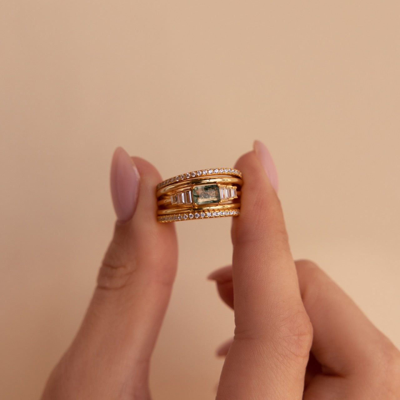 Moss agate ring stacked with three other gold bands, including pavé and domed styles, held between two fingers against a soft beige background.