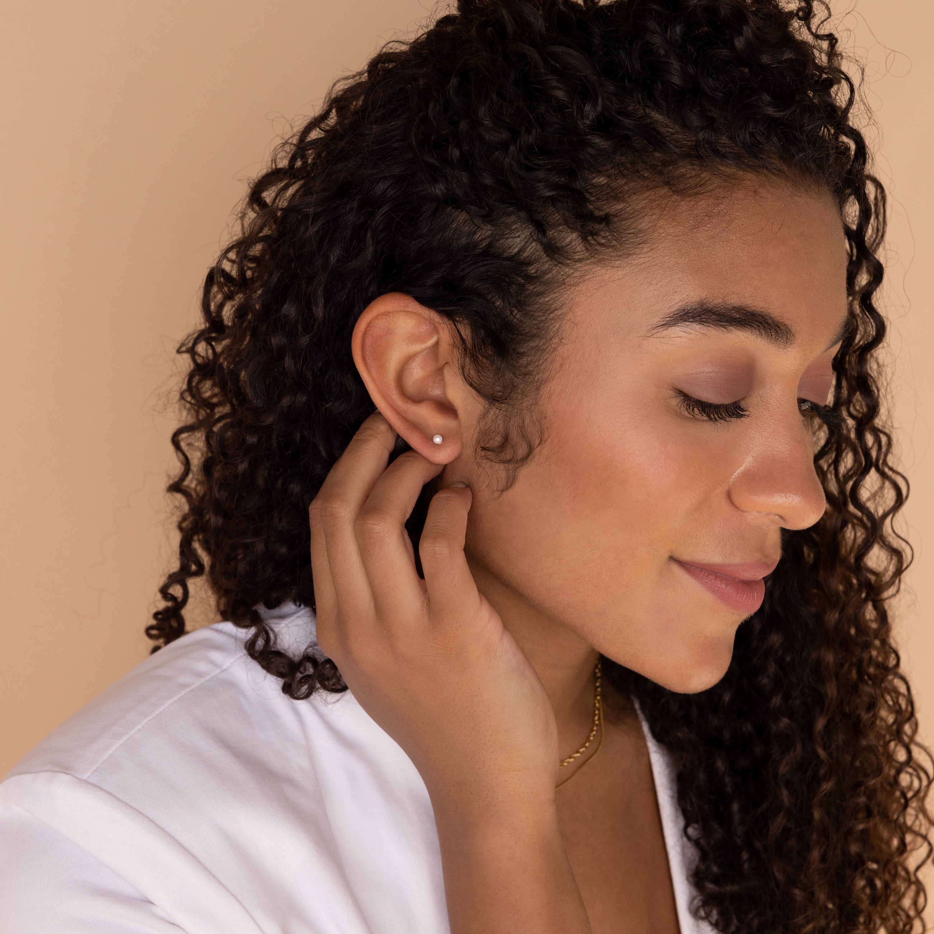A woman with curly hair touches her ear, wearing a white top and the Dainty Pearl Studs in Sterling Silver, against a beige background.