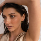 Woman with wet dark hair, wearing Shark Tooth Huggies in 18K Gold and a beige satin top, arm raised above her head—capturing effortless beach vacation vibes.