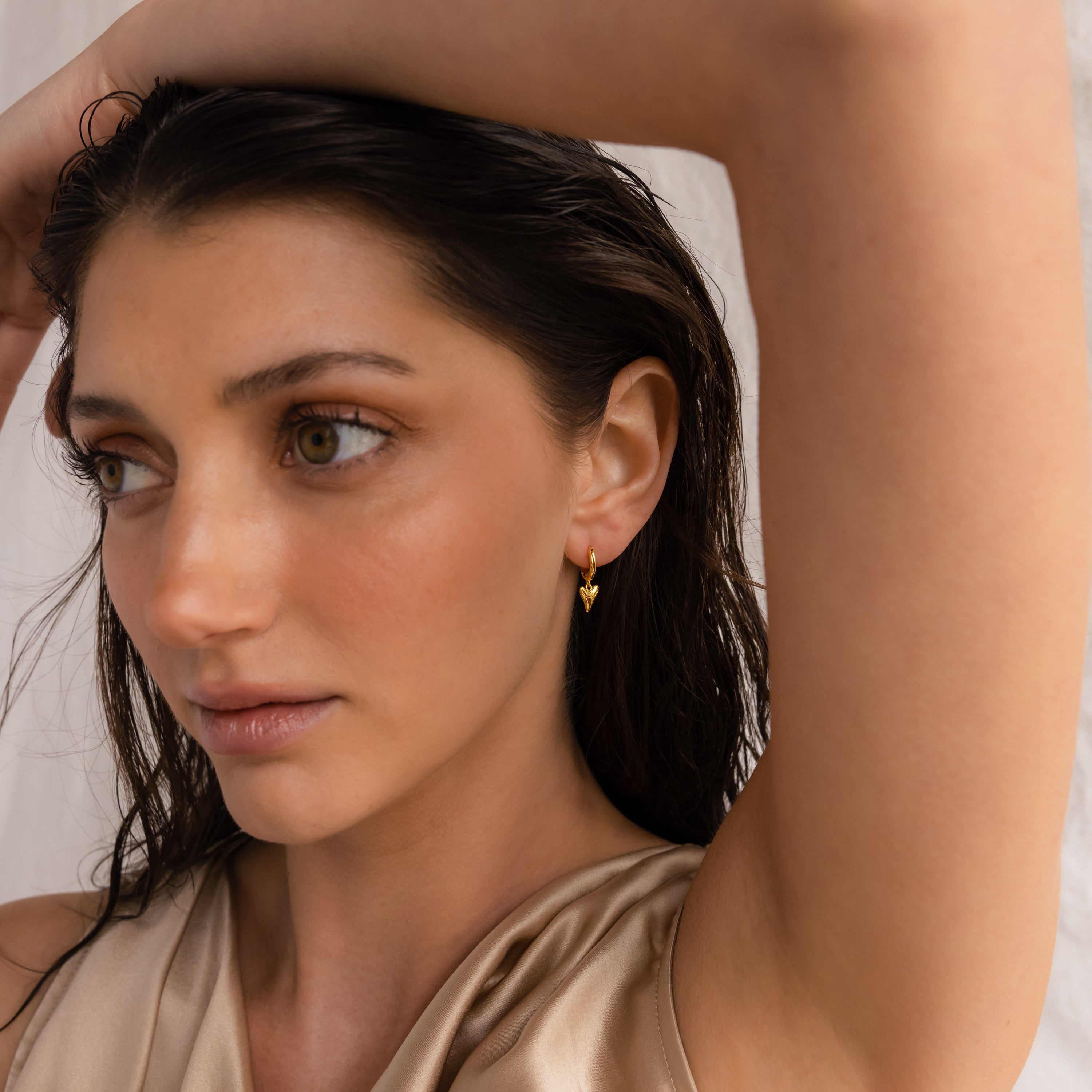 Woman with wet dark hair, wearing Shark Tooth Huggies in 18K Gold and a beige satin top, arm raised above her head—capturing effortless beach vacation vibes.