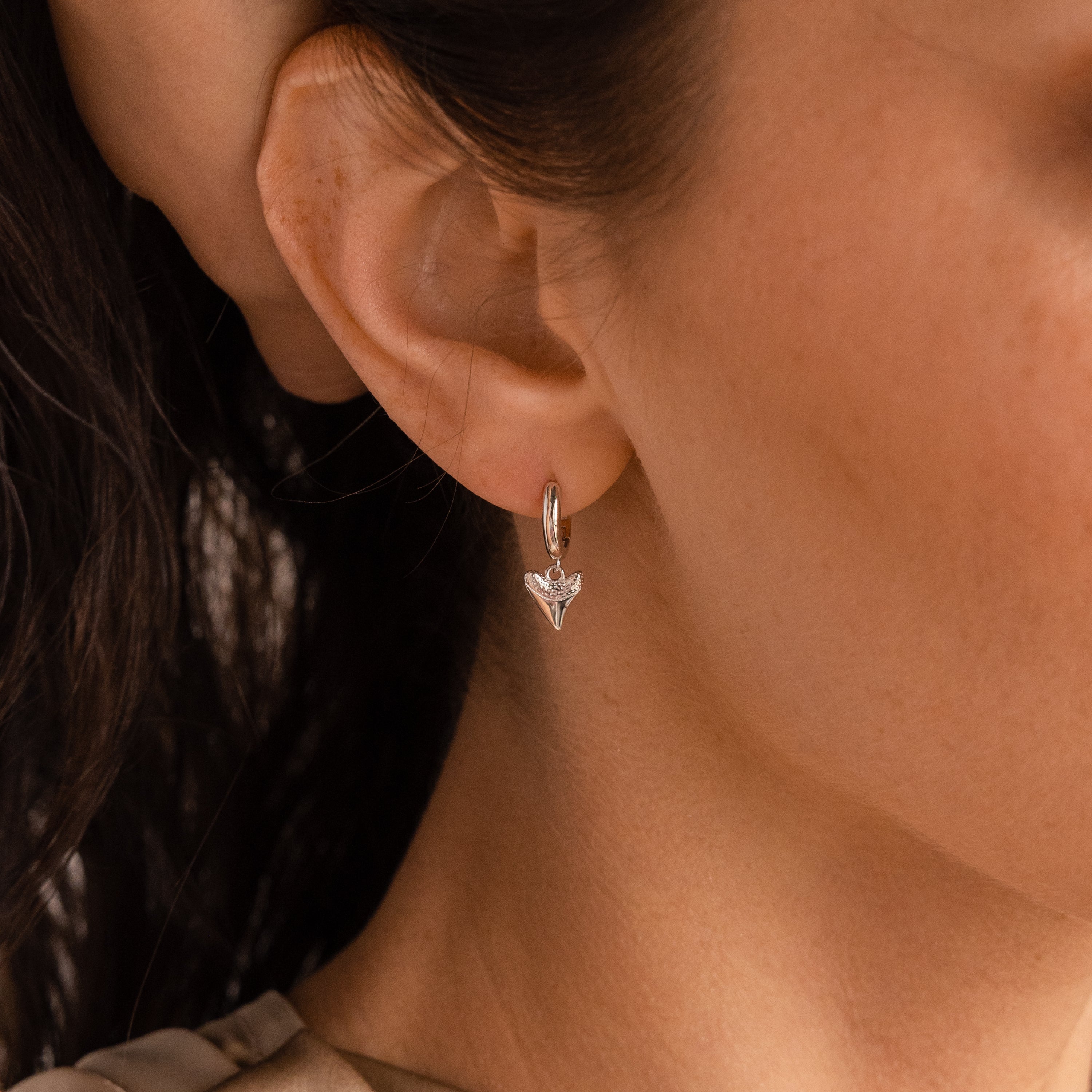 Close-up of a womans ear wearing Shark Tooth Huggies in Silver, small hoop earrings with a triangle-shaped pendant—perfect for adding coastal style to your beach vacation.