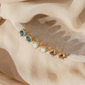 Six gold stud earrings, including the Halo Birthstone Studs, are arranged on a marble dish with a beige fabric backdrop.