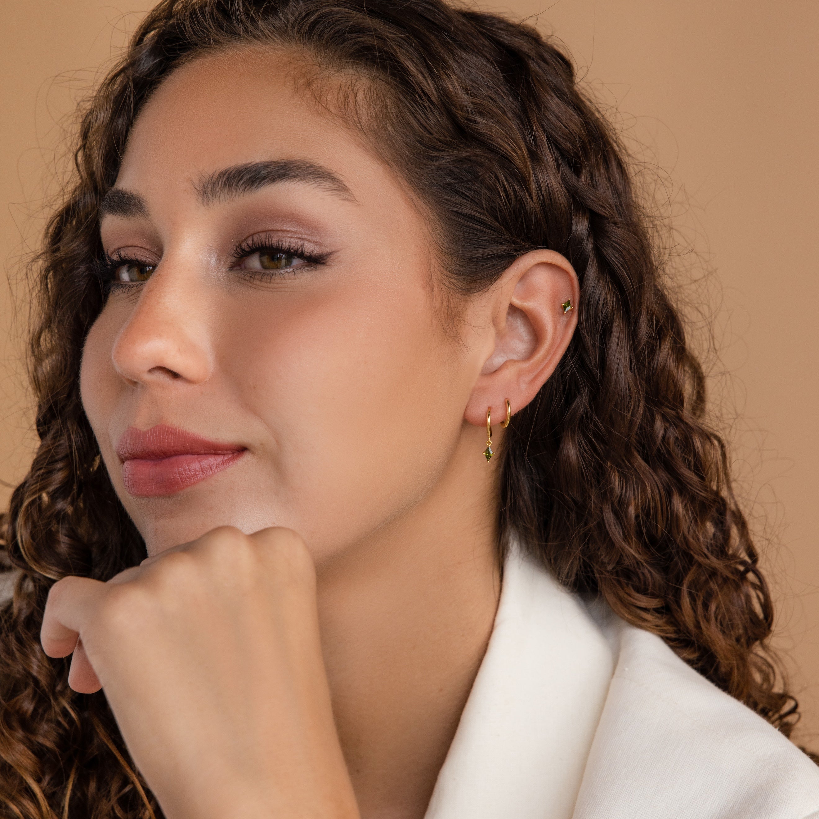 Woman with curly hair wearing Peridot Earrings Set in 18K Gold, resting her chin on her hand and gazing thoughtfully to the side.