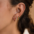 A close-up of an ear wearing three Vera Pave Emerald Huggies, each gold hoop adorned with vibrant red, blue, and green gemstones.