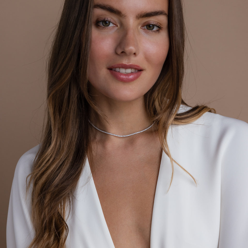 Woman wearing a deep V-neck white blouse and a sparkling diamond tennis choker necklace, smiling softly with loose, wavy hair against a neutral beige background.