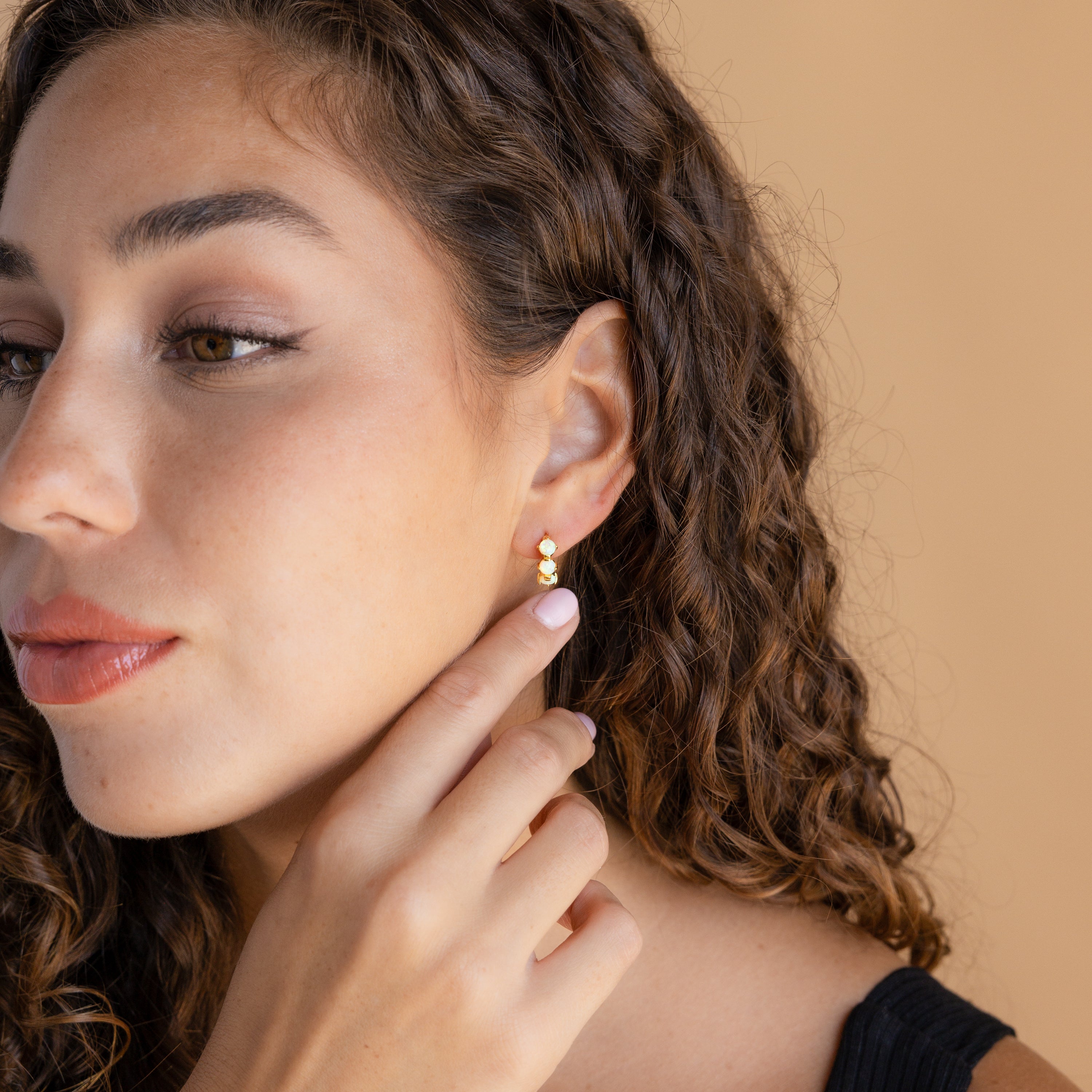 A woman with curly hair wears Triple Diamond Huggies, touching the elegant earrings as she gazes to the side against a beige background, highlighting the minimalist beauty of this jewelry.