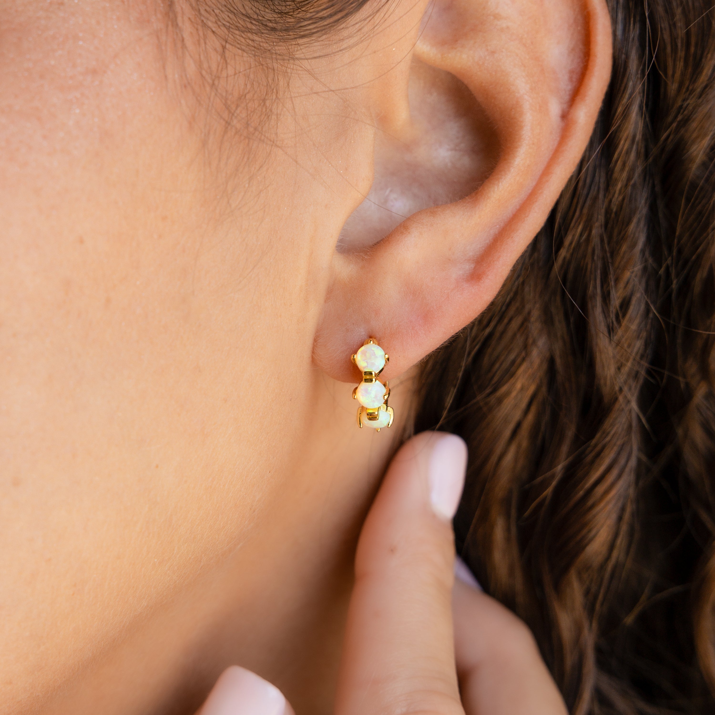 A woman's ear adorned with a double opal stud earring, her hand gently touching her earlobe—beautifully complemented by the personalized sparkle of the Triple Birthstone Huggies.