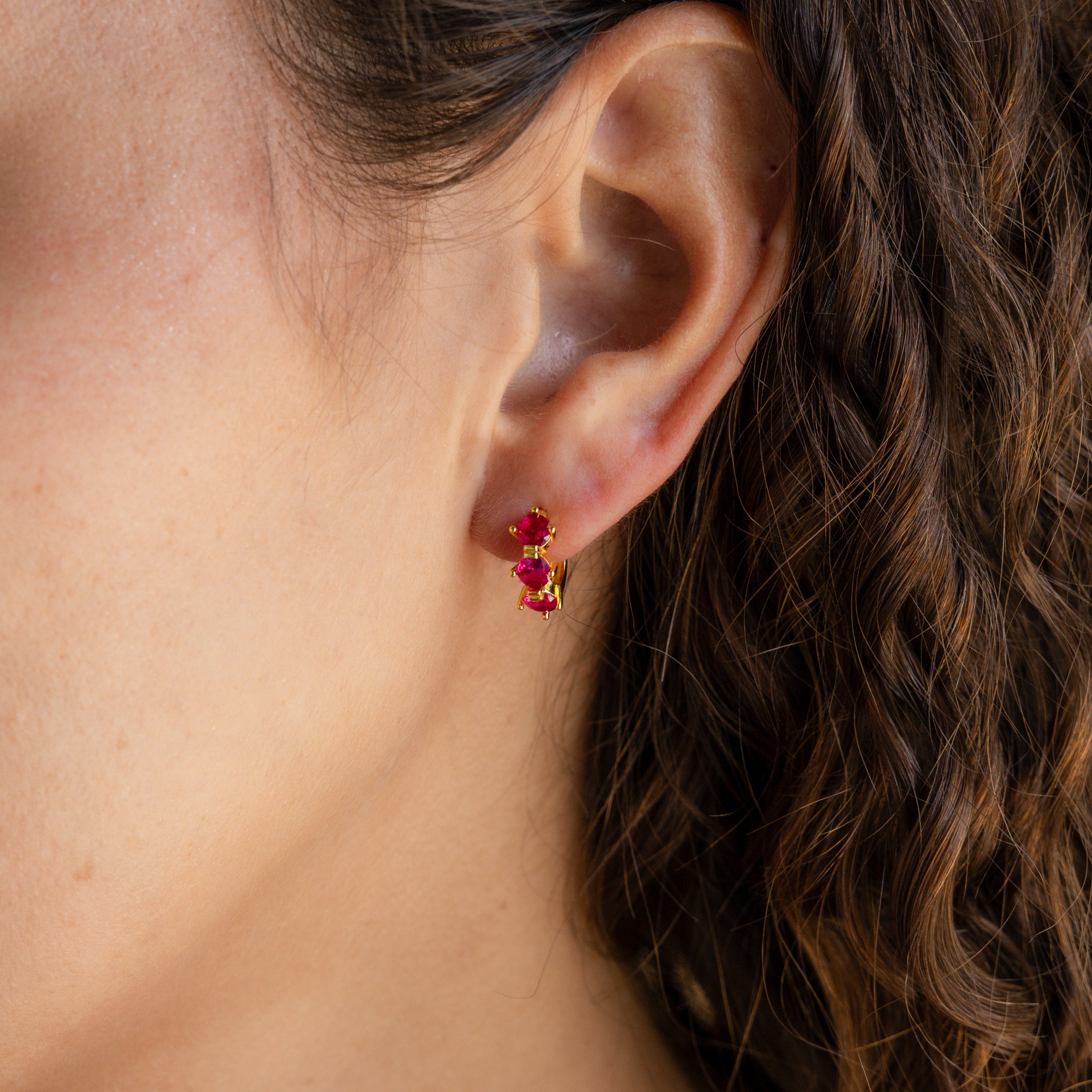 Close-up of a woman's ear with Triple Ruby Birthstone Huggies in gold and wavy brown hair.
