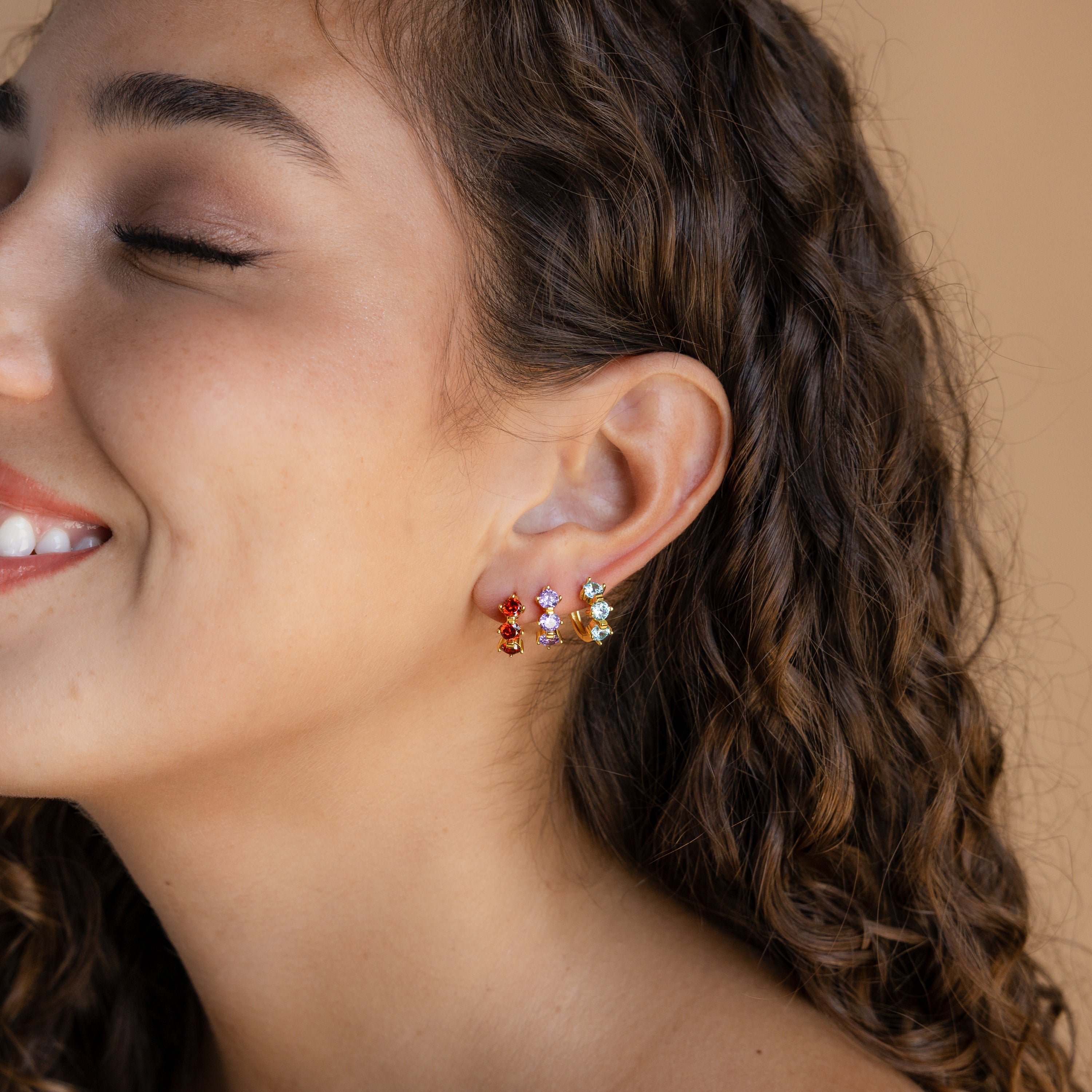 A woman with curly hair smiles, wearing vibrant Triple Blue Zircon Huggies for a subtle pop of color. These gemstone earrings pair perfectly with minimalist hoops. The background is beige.