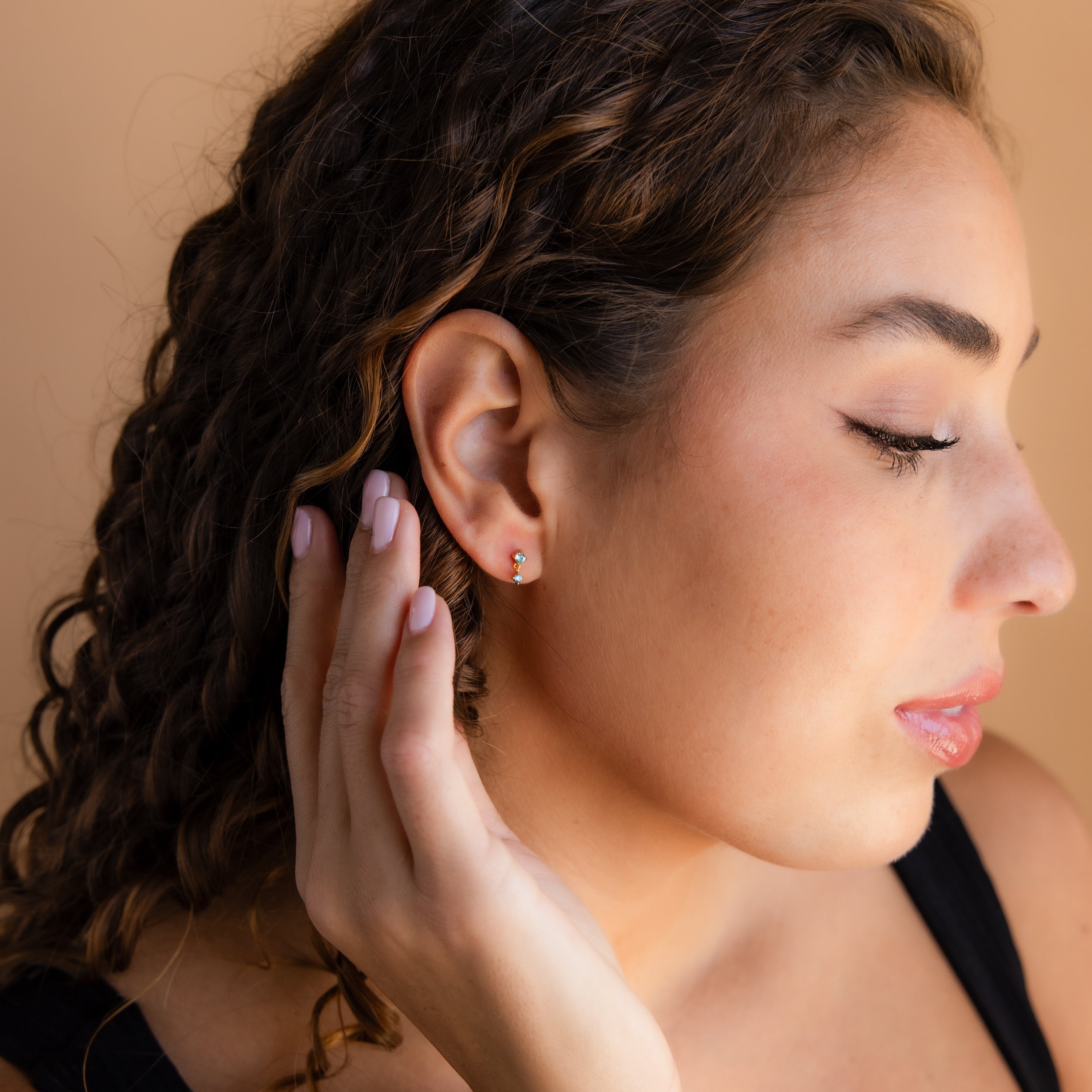 Woman with wavy brown hair, eyes closed, touches her accessory while wearing a black top and elegant Sapphire Drop Studs against a beige background.