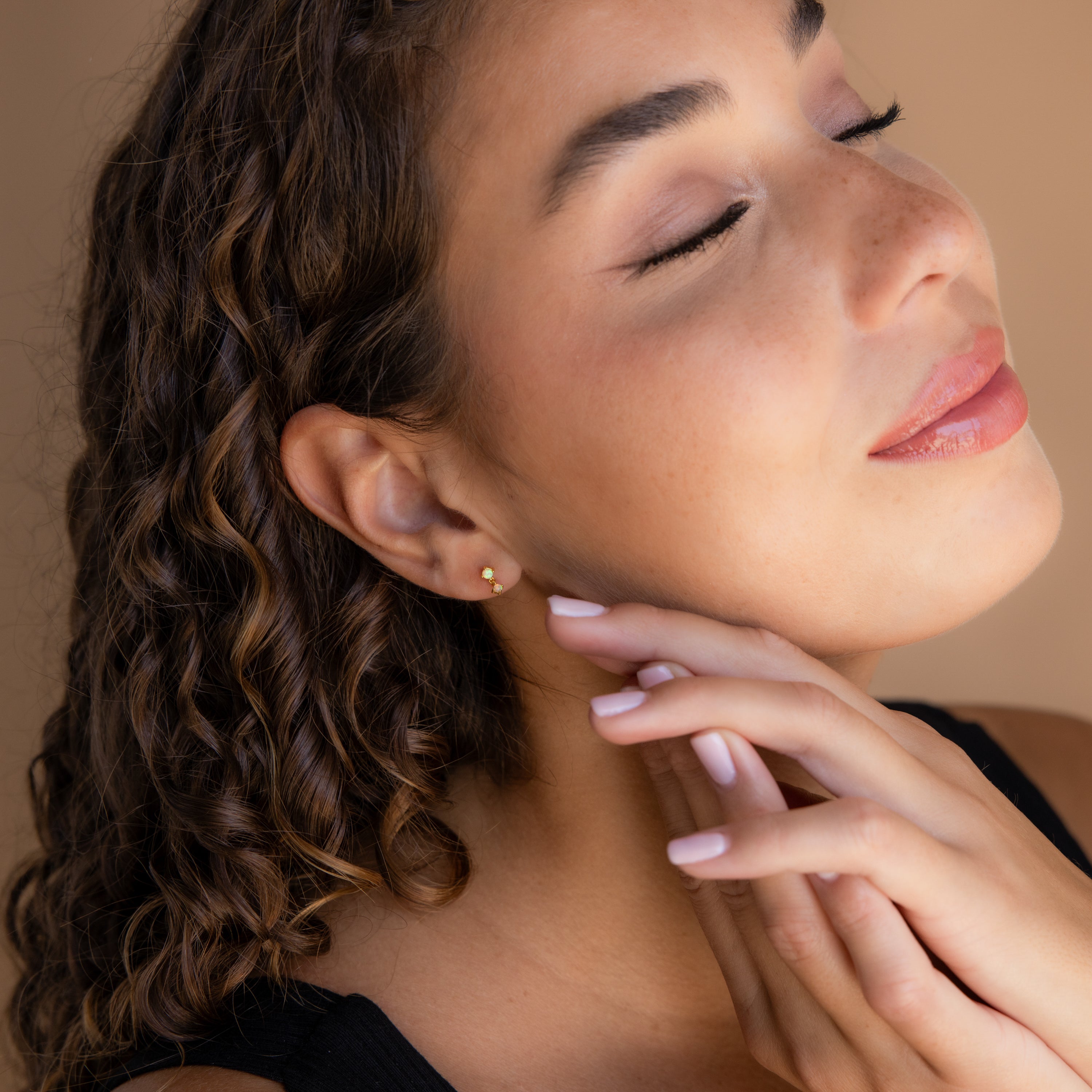 A woman with curly hair and closed eyes gently touches her neck, smiling softly against a beige background while wearing elegant Opal Drop Studs as a delicate accessory.