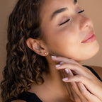A woman with curly hair and closed eyes touches her face, her peaceful expression enhanced by the elegant Diamond Drop Studs she wears.