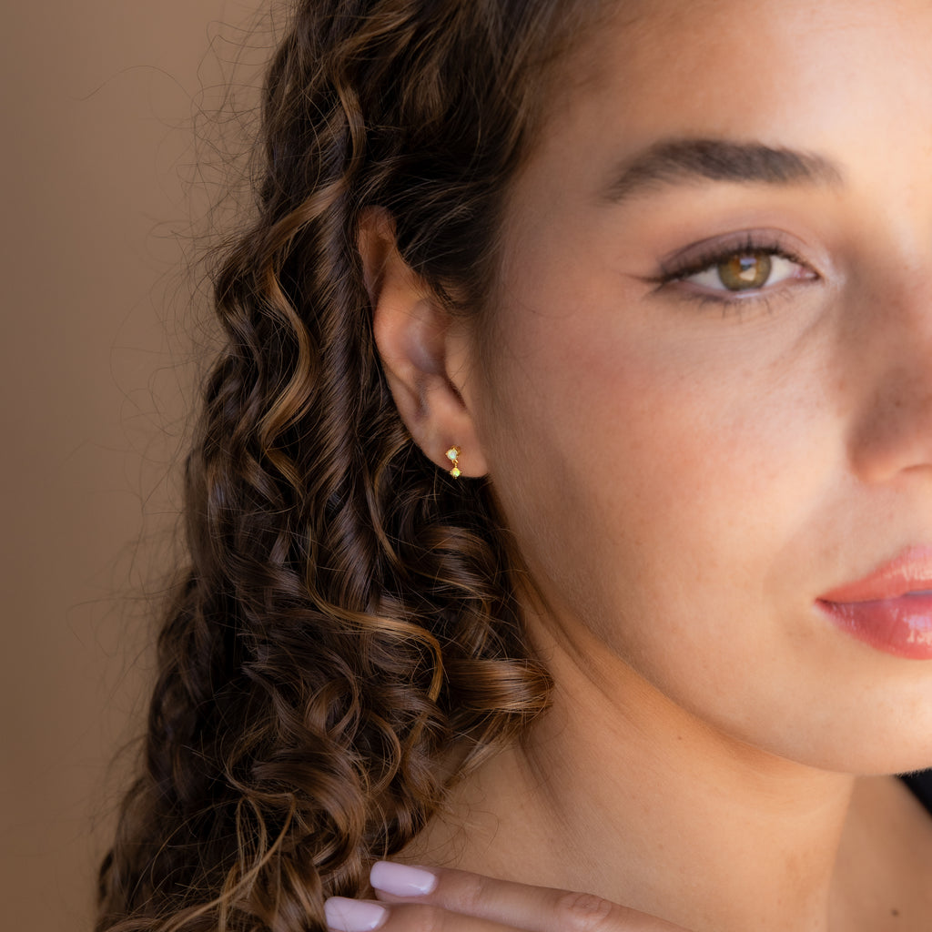Woman with curly hair wearing Birthstone Drop Studs and a small gold hoop earring, gently touching her neck and gazing ahead.