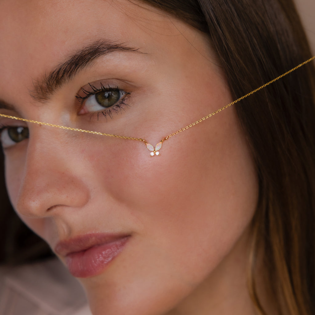 Close-up of a woman’s face with the Pink Opal Butterfly Necklace, a delicate gold pendant featuring a small butterfly charm, held across her cheek.