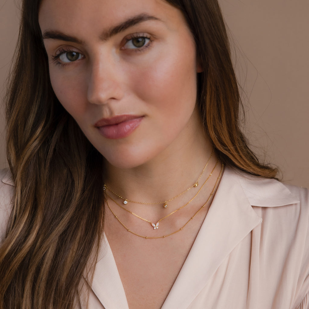 Woman with long brown hair wears a Pink Opal Butterfly Necklace and a pale pink blouse, posing against a beige background.