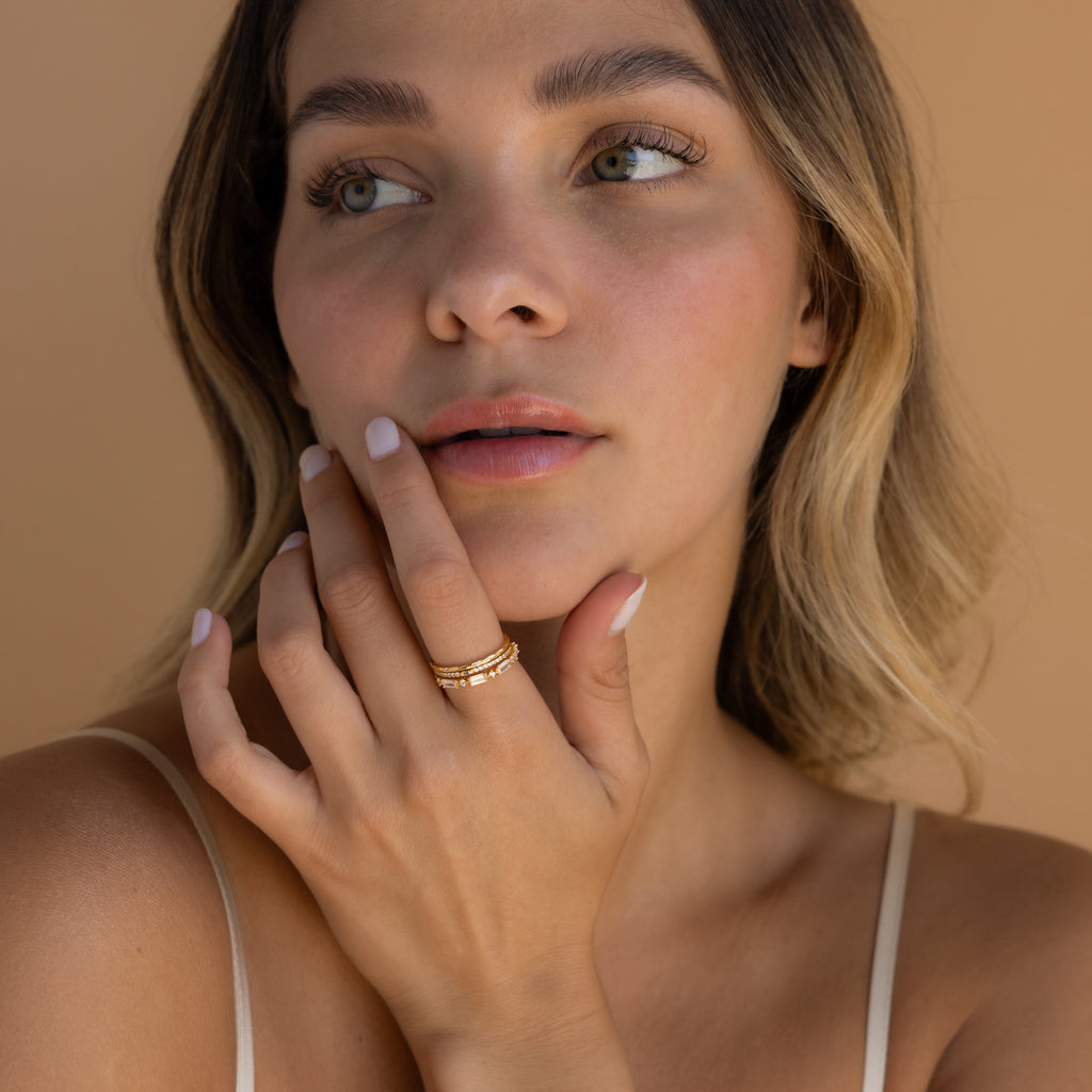 Woman with wavy hair touches her face, wearing the Diamond Stacking Ring Set in 18K Gold and a beige spaghetti strap top against a tan background—perfect Christmas gift inspiration.