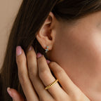 Close-up of a model wearing gold Aquamarine Diamond Huggie earrings, featuring three sparkling blue stones, paired with a gold twisted ring on her hand touching her face.