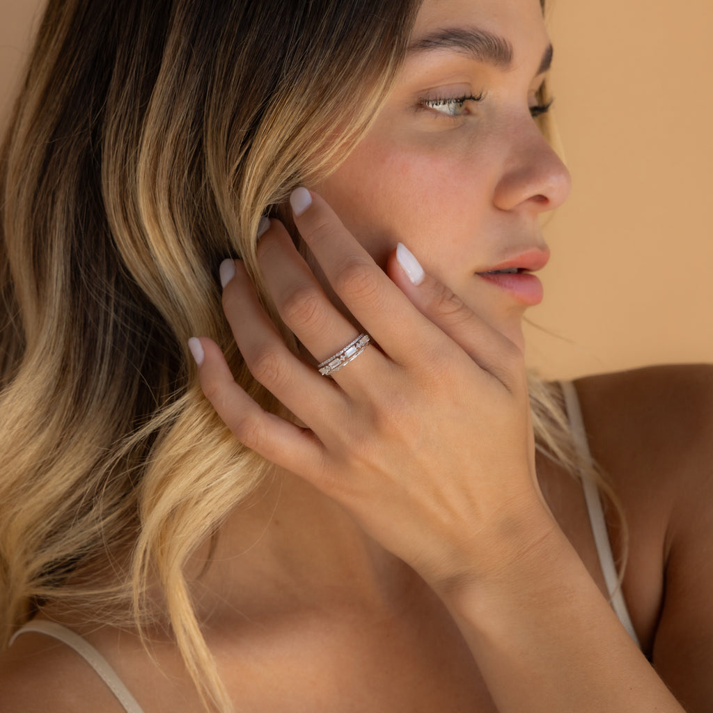 A woman with blonde hair gently touches her face, displaying the Diamond Stacking Ring Set on her finger against a beige background—a perfect Christmas gift for someone special.