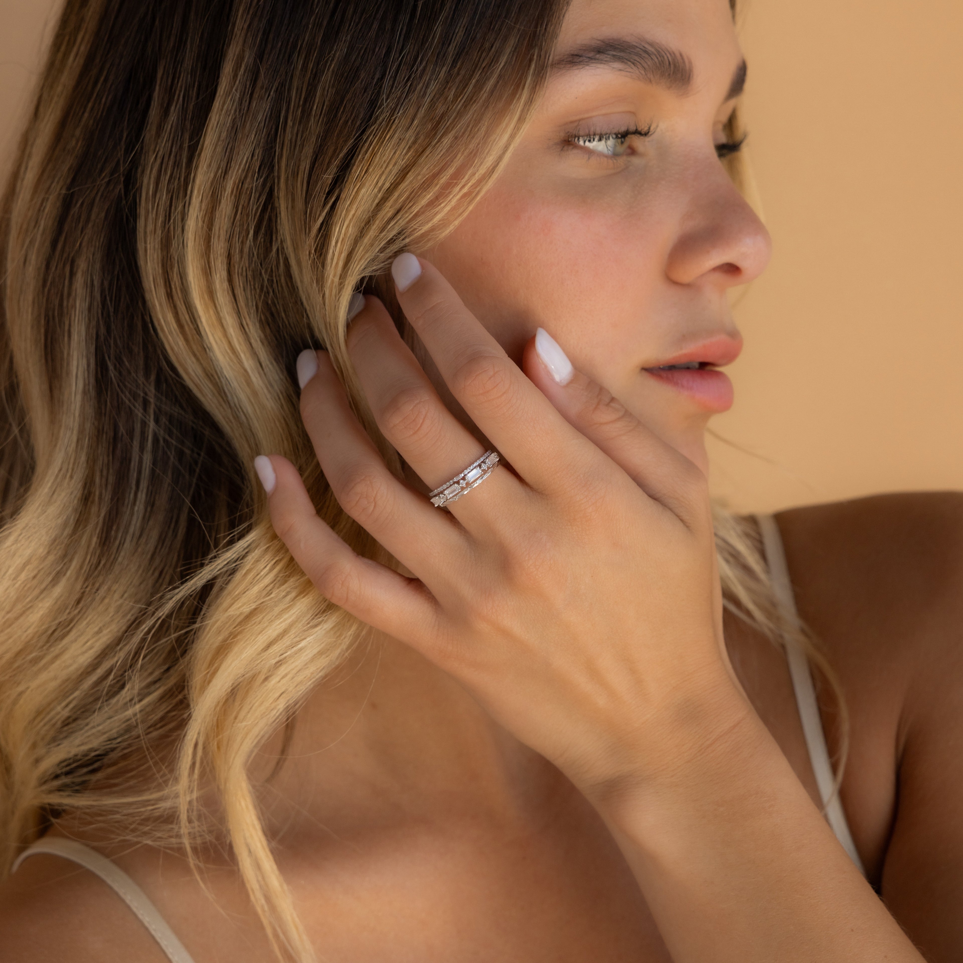 A woman with blonde hair gently touches her face, displaying the Diamond Stacking Ring Set on her finger against a beige background—a perfect Christmas gift for someone special.