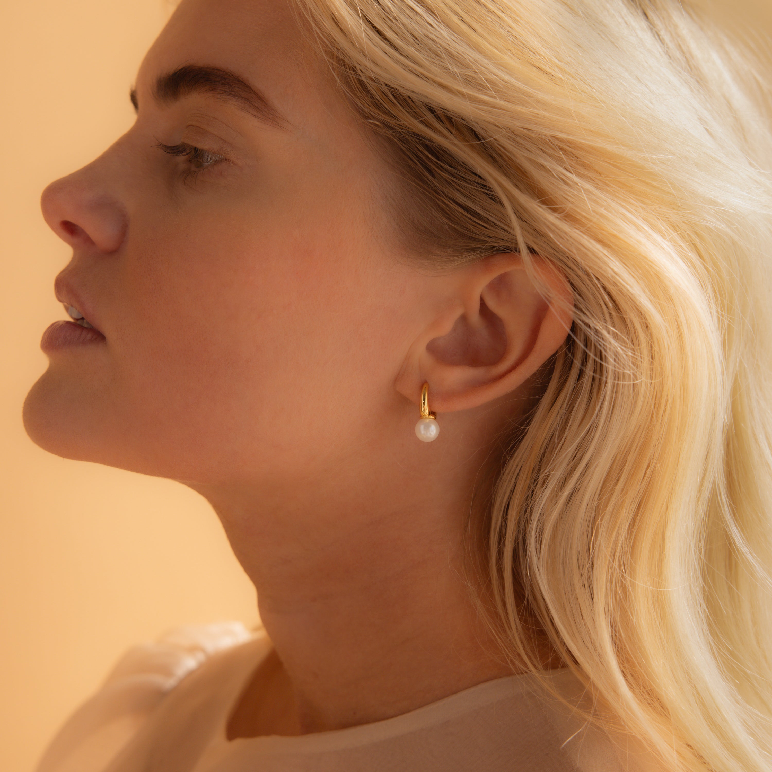 A gold pearl drop huggie earring worn on a model's ear, showcasing its minimalist and timeless design