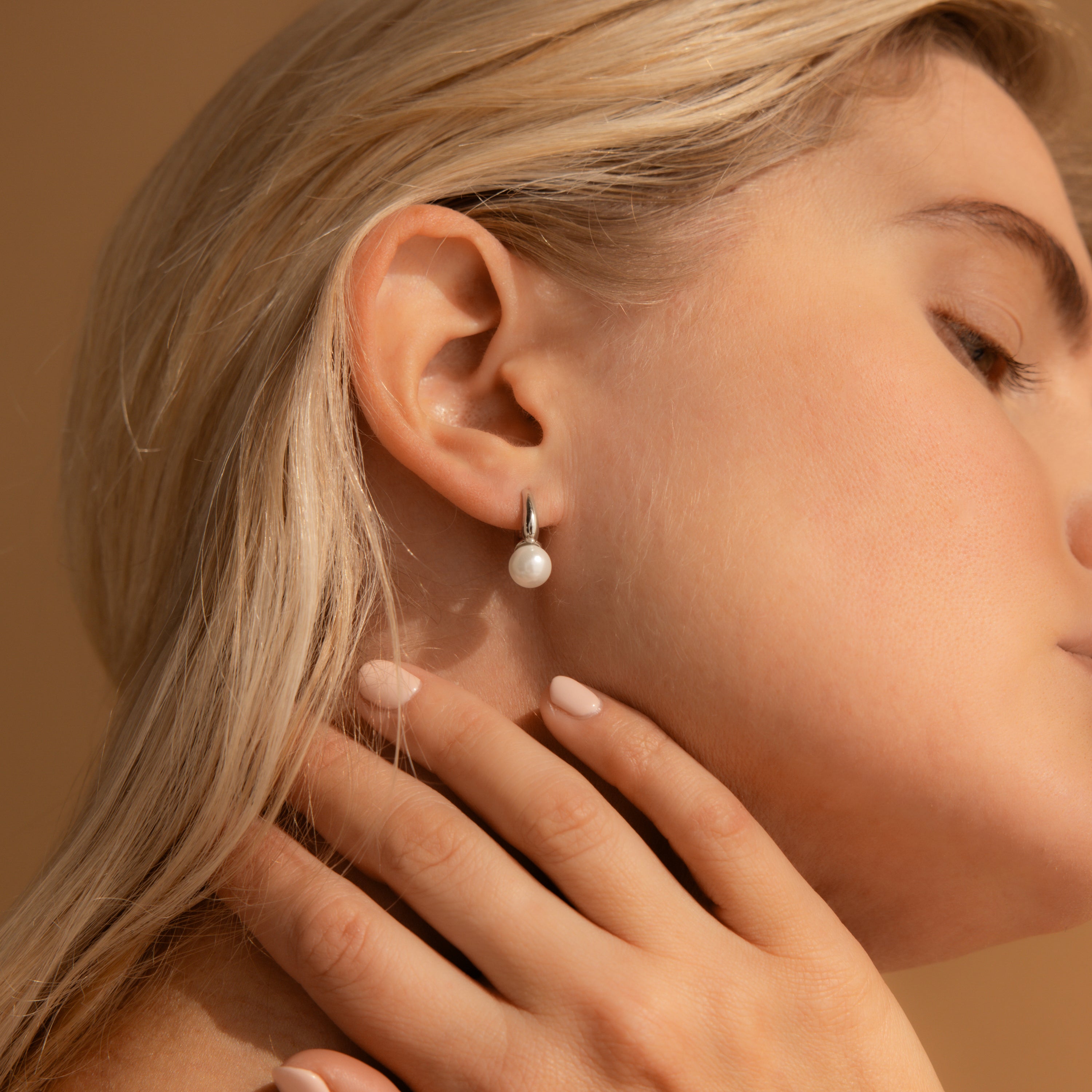 A woman wearing a silver huggie earring with a single white pearl drop, shown close-up on her ear with her hand gently touching her neck