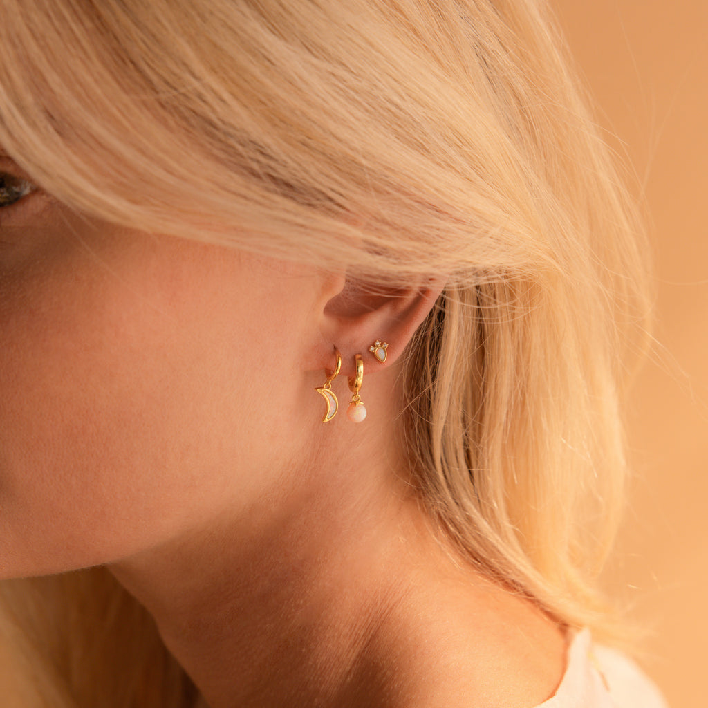 Close-up of a woman’s ear adorned with the Opal Drop Earrings Set—featuring a moon, opal drop, and small stud—highlighting effortless earring layering against a beige background.