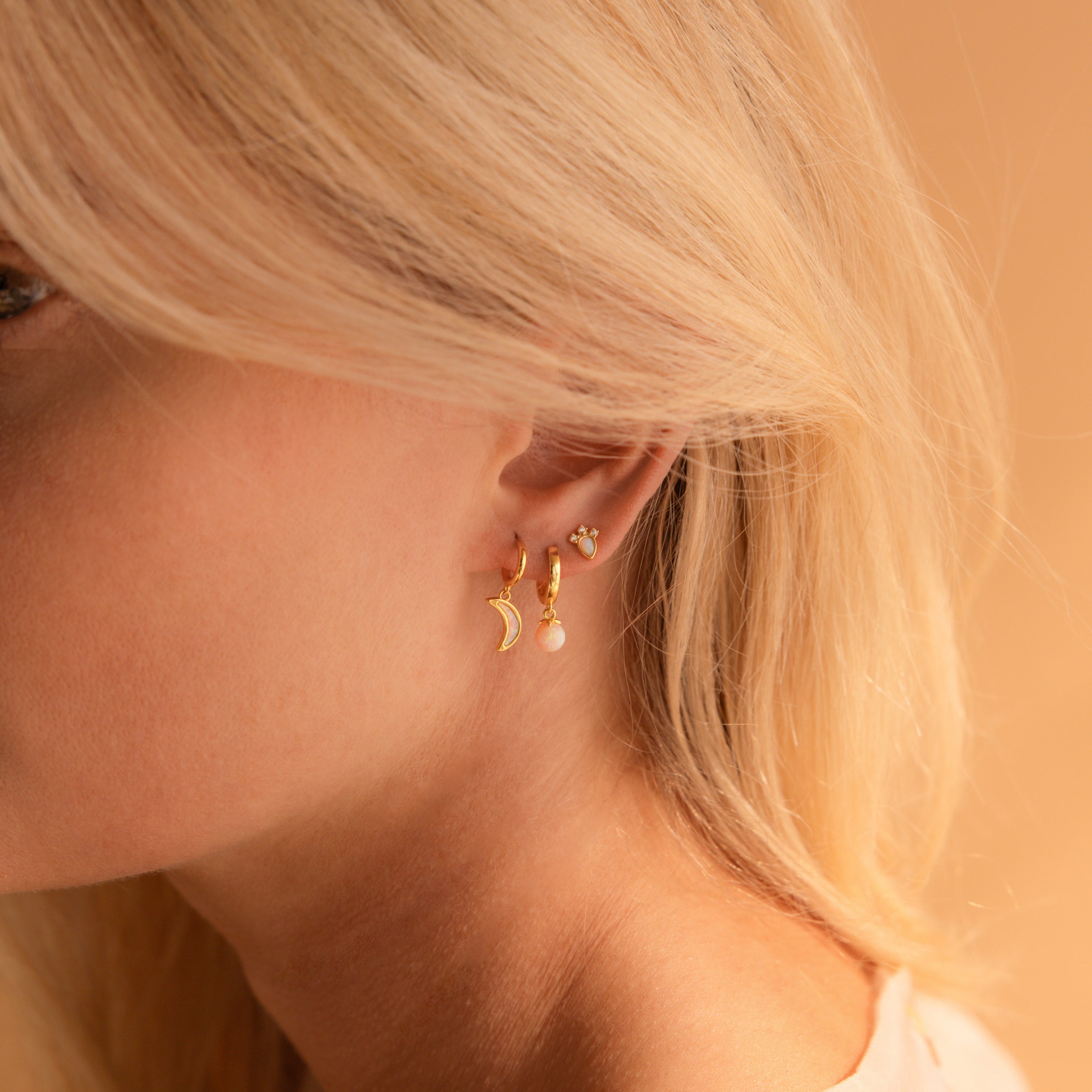 Close-up of a woman’s ear adorned with the Opal Drop Earrings Set—featuring a moon, opal drop, and small stud—highlighting effortless earring layering against a beige background.