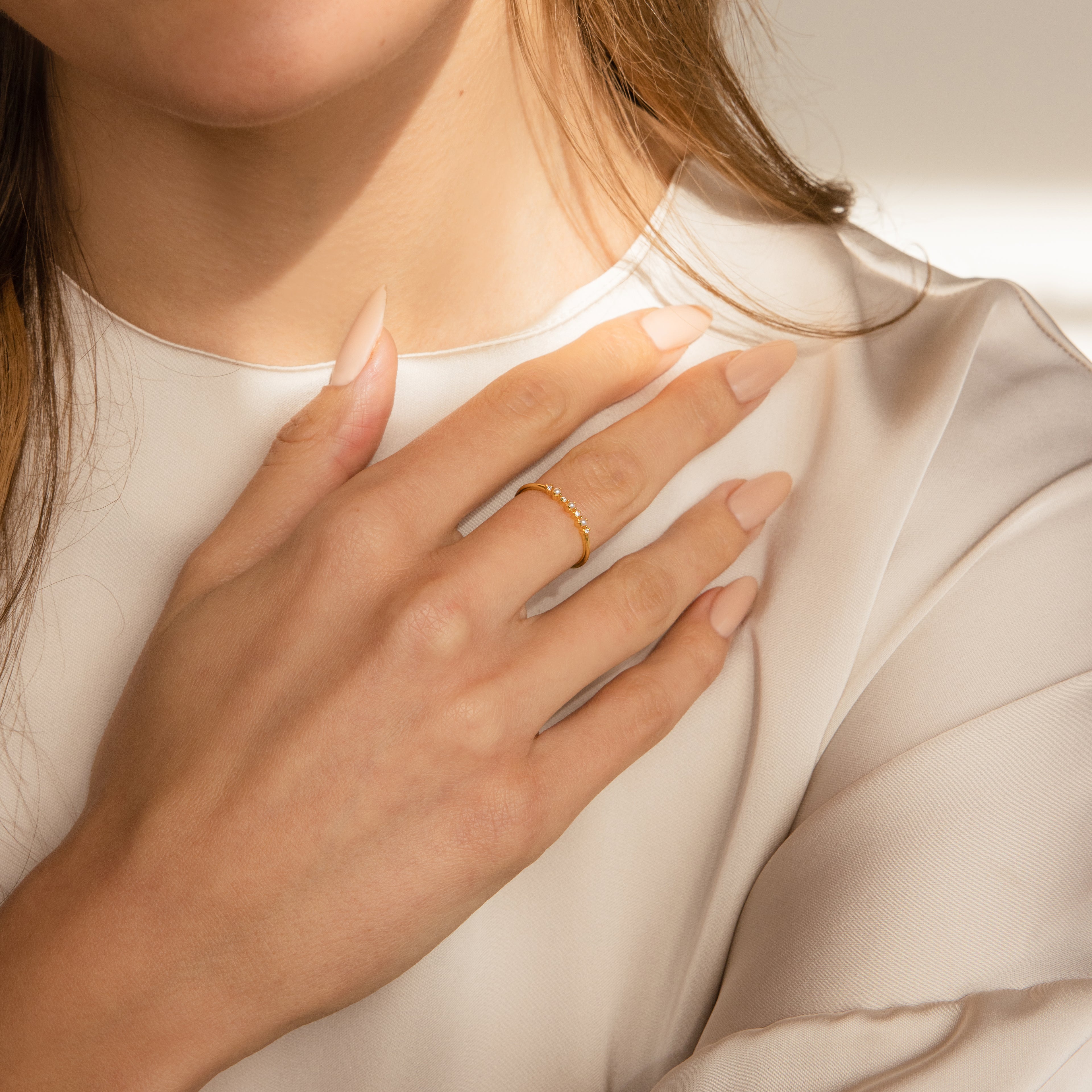A woman with long nude nails wears the Delicate Pearl Diamond Ring, gently touching her chest over a cream-colored blouse.