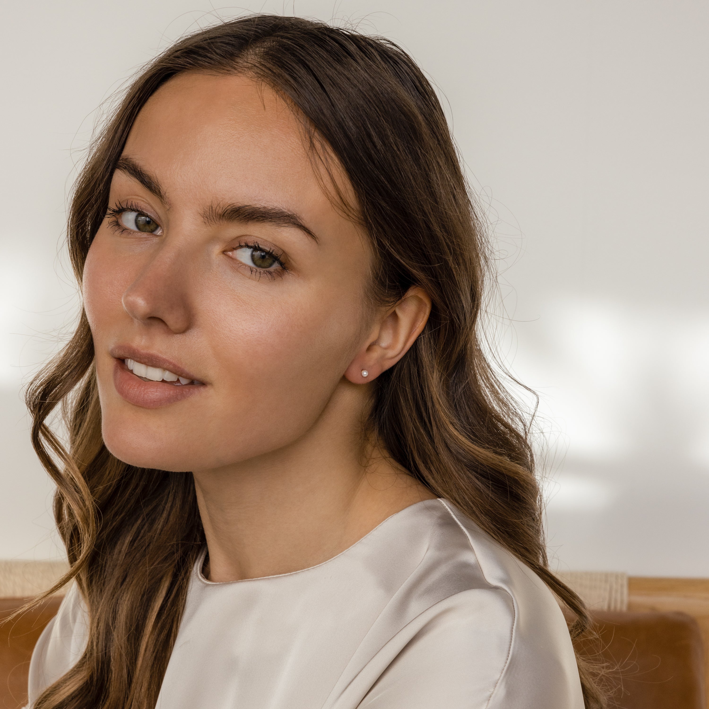 Woman with long brown hair and a light satin top smiles gently indoors under soft lighting, wearing Dainty Pearl Studs that add a subtle touch of elegance to her look.