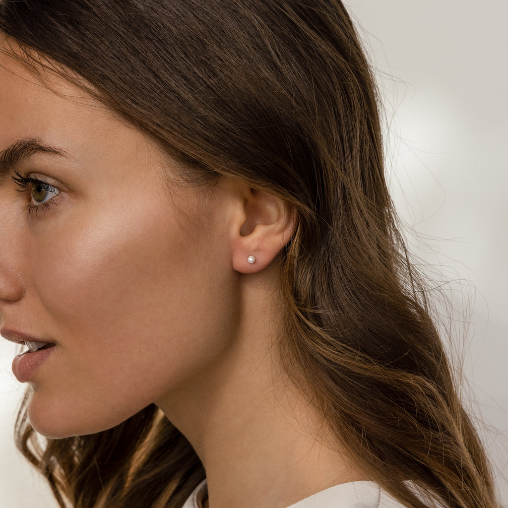 Woman with wavy brown hair wears Dainty Pearl Studs in Sterling Silver, shown in profile against a soft background.