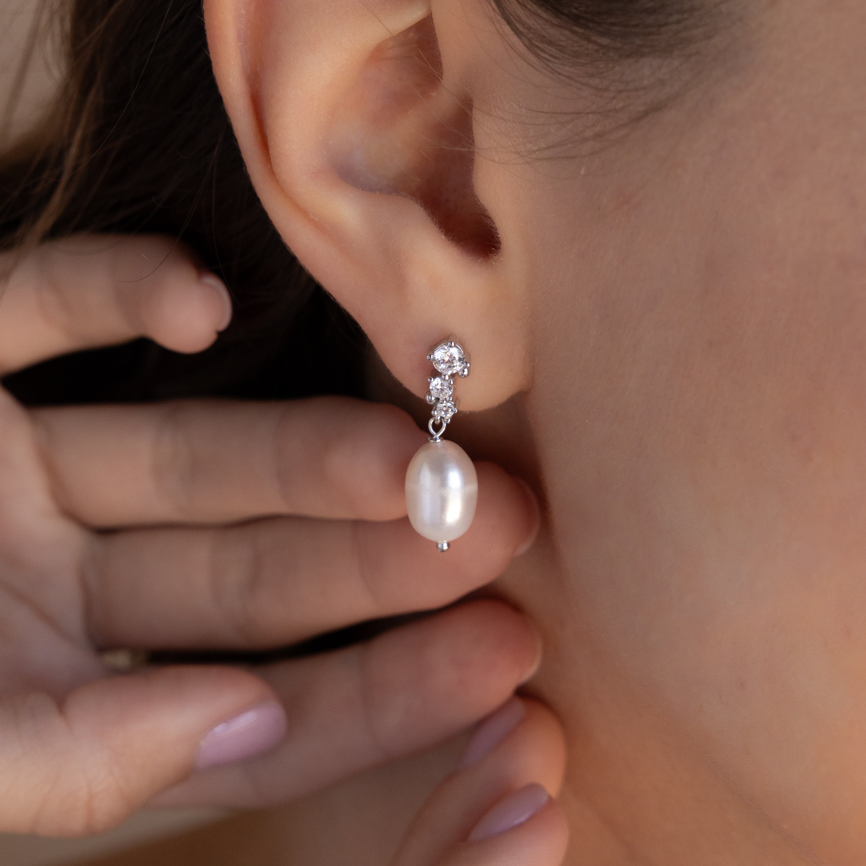 Close-up of a silver Millie Drop Earring worn on the ear, featuring three small round white stones and a dangling oval pearl.