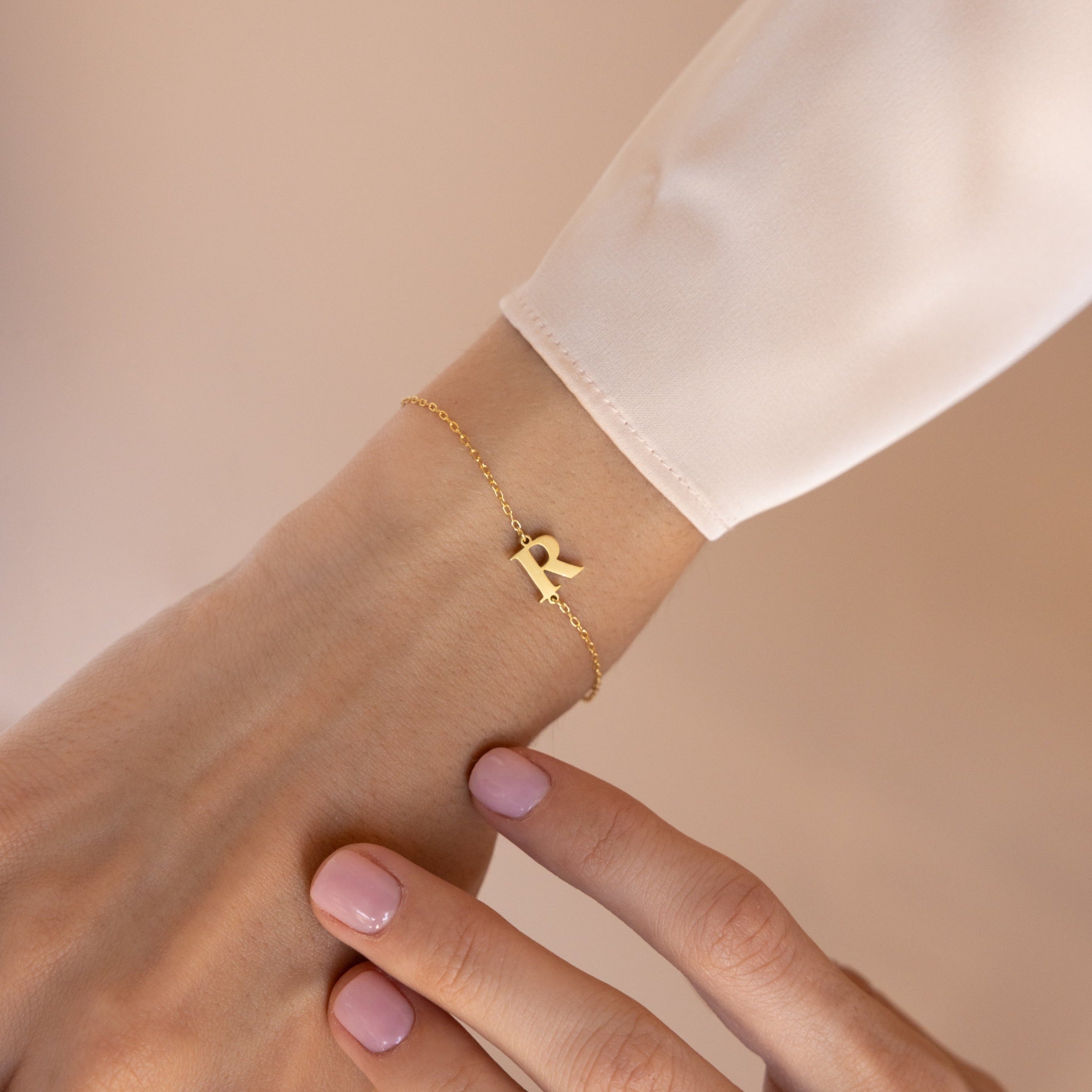 The Newsletter Initial Bracelet, featuring a gold R charm, elegantly adorns the wrist as the other hand gently touches this personalized accessory.