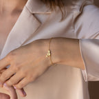 The Carrie Initial Bracelet, featuring a letter A charm, elegantly adorns the wrist when styled with a light beige satin blouse—a refined take on personalized jewelry.