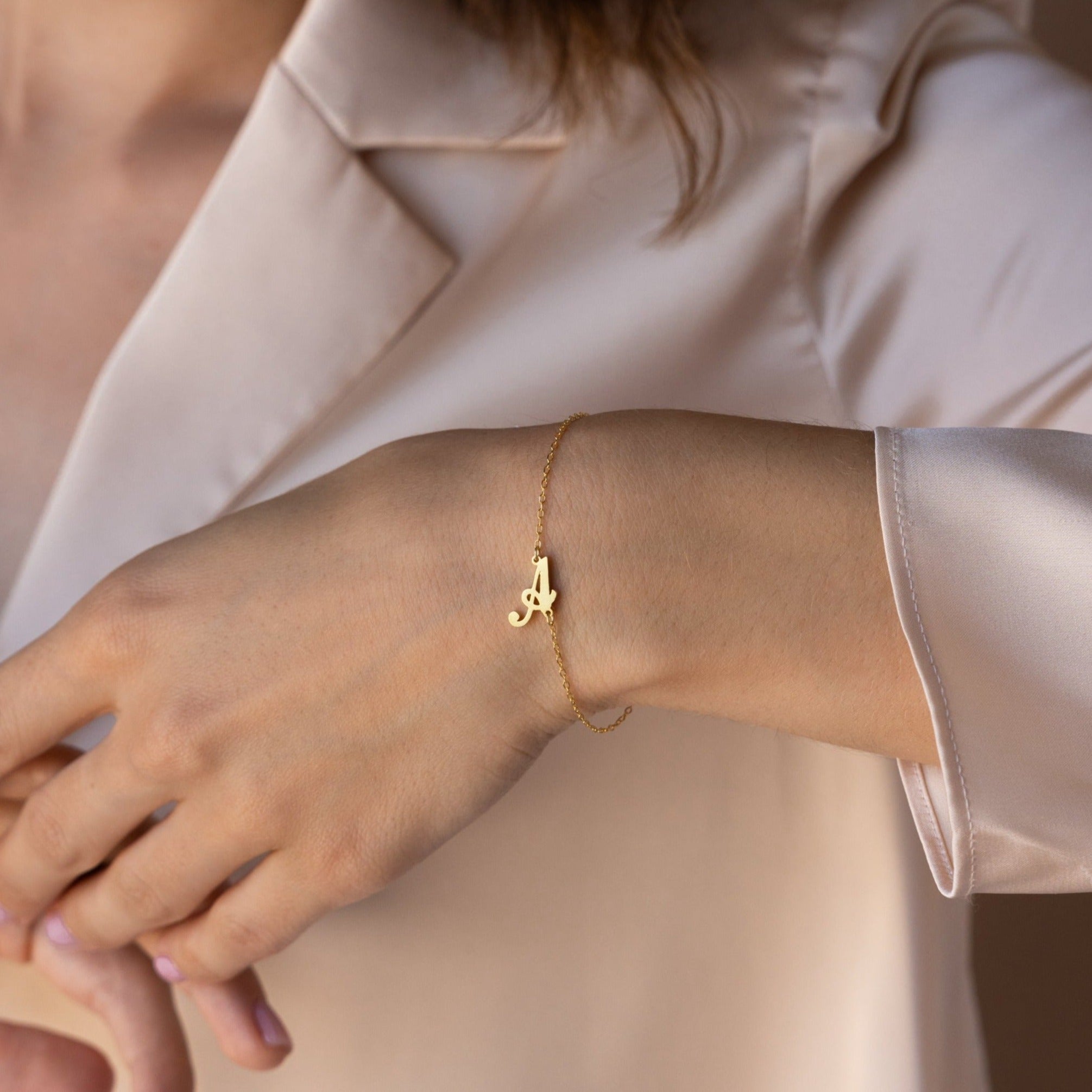 The Carrie Initial Bracelet, featuring a letter A charm, elegantly adorns the wrist when styled with a light beige satin blouse—a refined take on personalized jewelry.