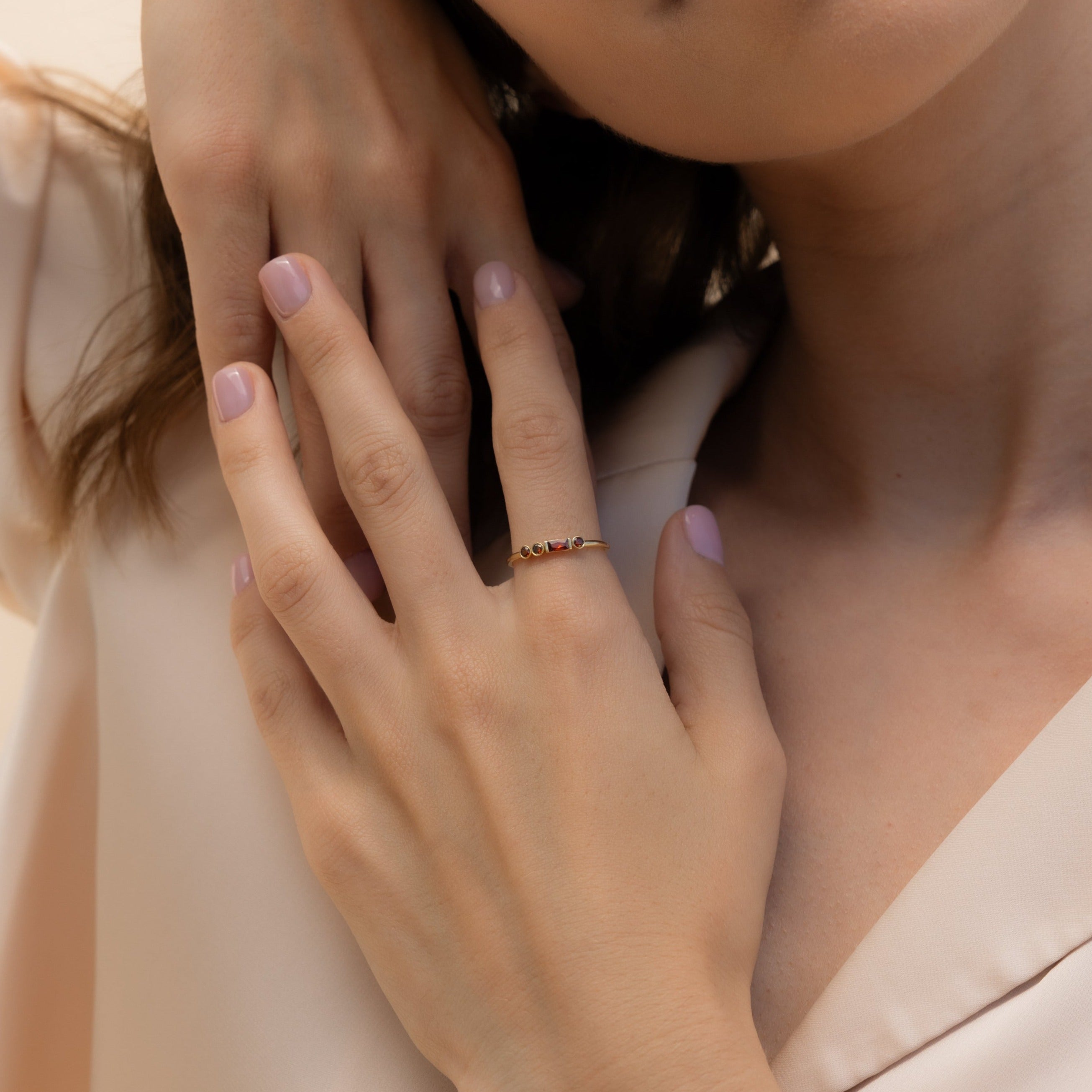 A woman with light pink nails touches her collarbone, highlighting a delicate gold Morse Code Birthstone Ring on her finger.
