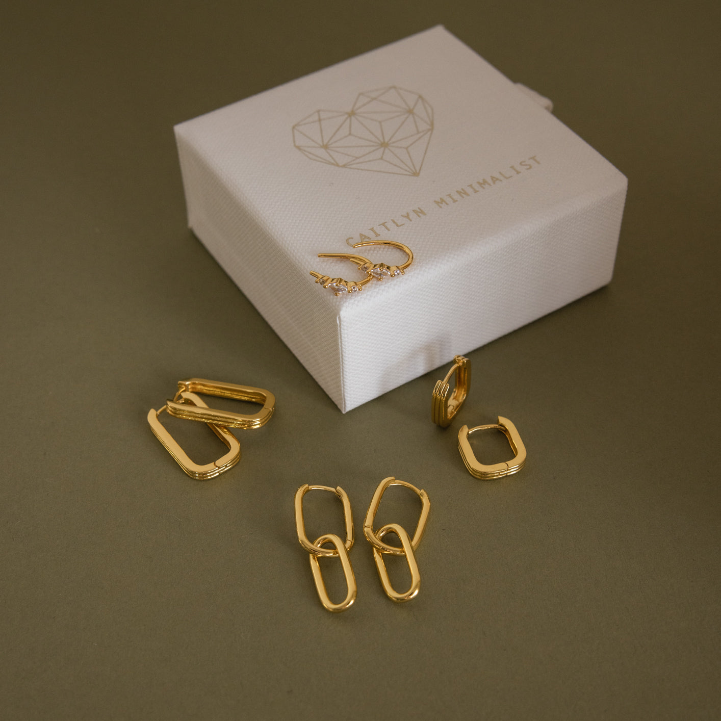 Monthly Earrings Subscription Box featuring minimalist new earrings in elegant 18K Gold, displayed beside a white jewelry box adorned with a geometric heart design.