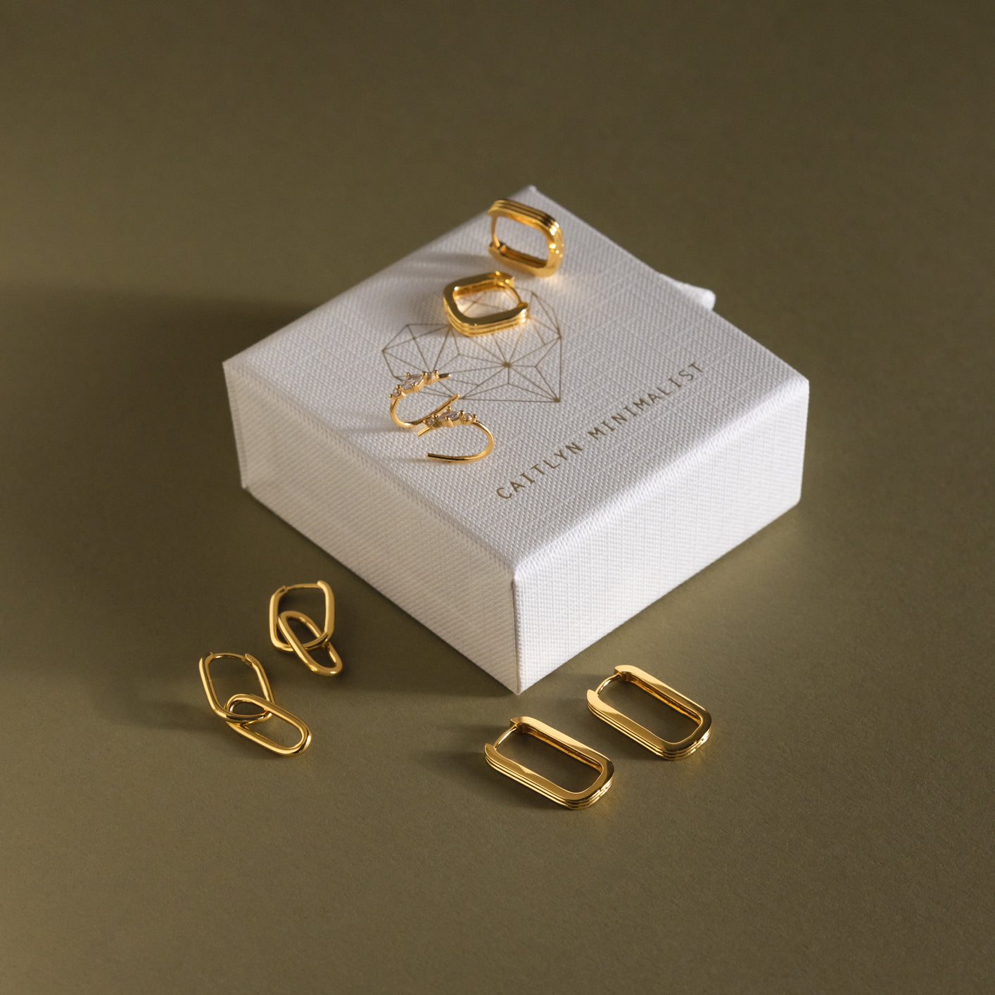 18K gold geometric earrings displayed by a textured white jewelry box on an olive background—find your next favorite pair with our Monthly Earrings Subscription Box.
