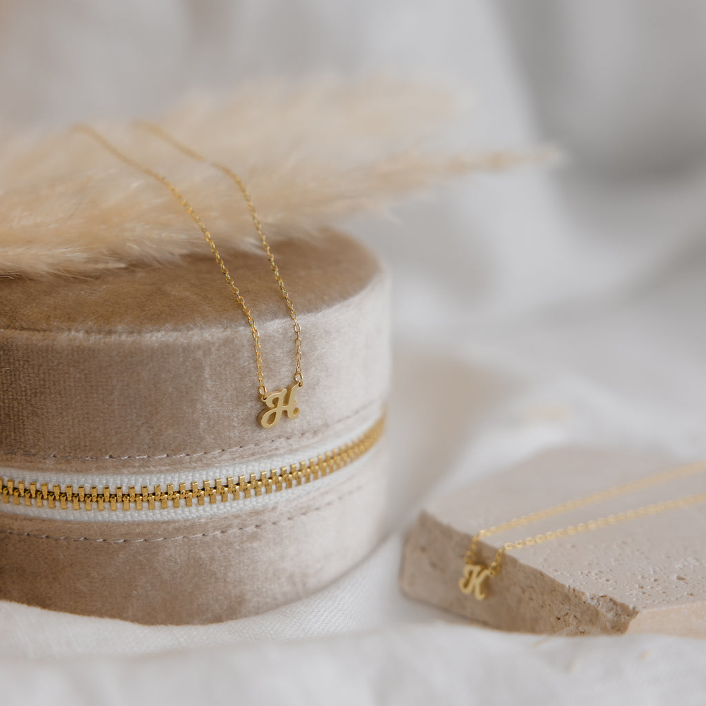 The Mellow Dainty Initial Necklace with a gold letter K pendant rests on a beige velvet jewelry box, next to another custom accessory displayed on a stone surface.
