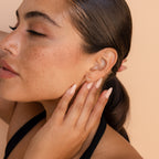 A close-up lifestyle shot of a woman with her hair pulled back, wearing a our diamond climber earring. She is touching her cheek with her hand, set against a solid warm beige background.